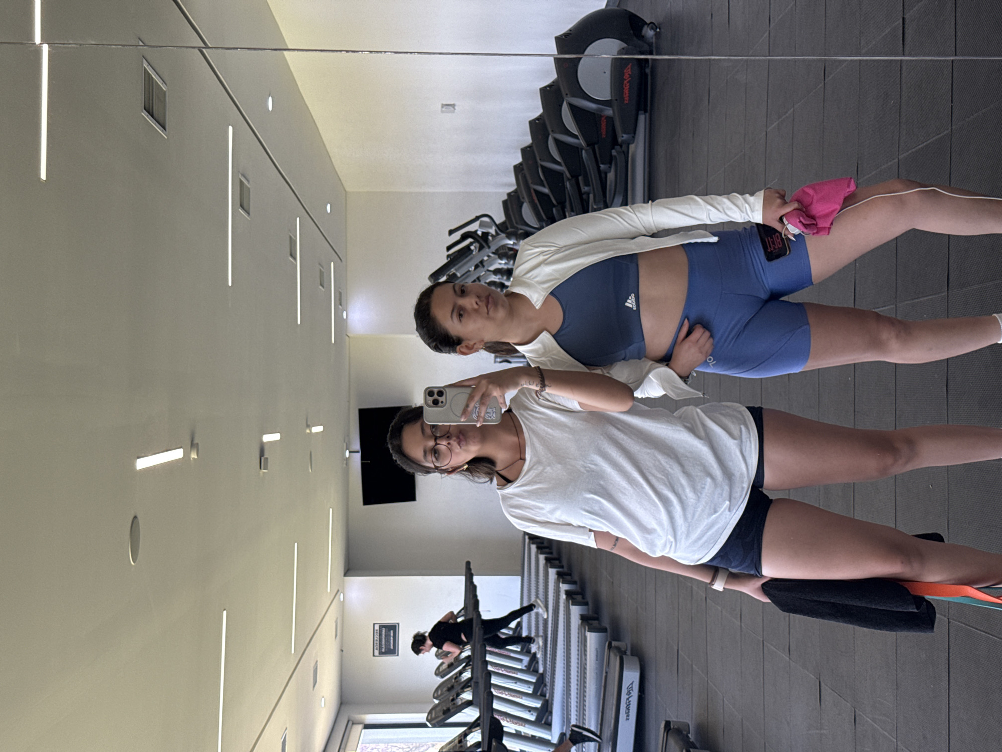 Two women are posing for a photo in a gym in San Andrés Cholula, Mexico. The woman in the foreground is holding a cell phone and wearing a white t-shirt, navy blue shorts, and glasses. She is standing next to a mirror, in which a reflection of the second woman can be seen. The second woman is wearing a white jacket, a navy blue sports bra, and blue shorts. In the background, there are treadmills and stationary bikes. Someone is on one of the treadmills.