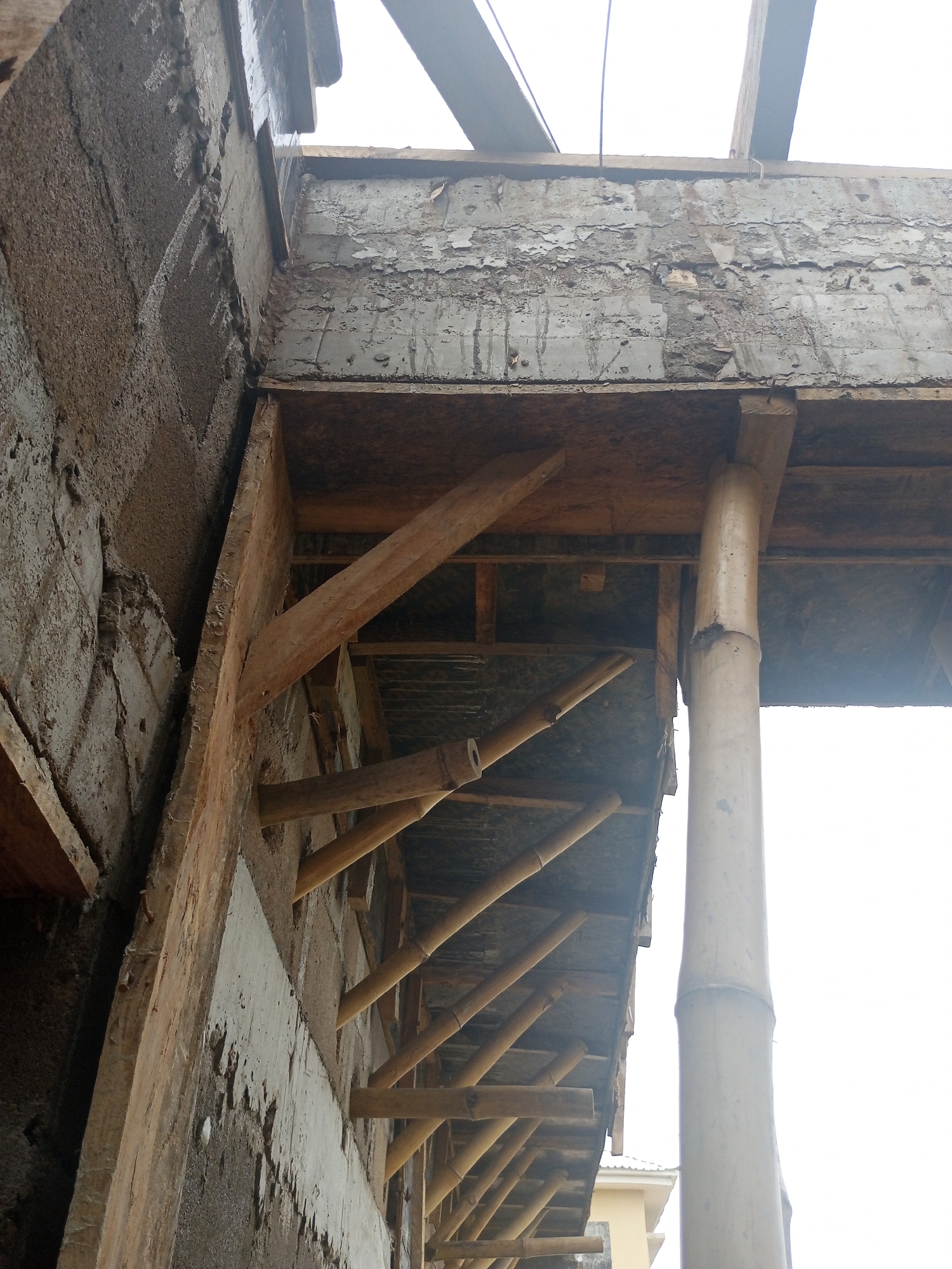 The image depicts an under-construction building with visible wooden and bamboo structural supports. A large, thick bamboo pole acts as a vertical support. Several diagonal bamboo poles are angled upwards, suggesting they are part of a roof or balcony structure. Wooden beams are also integrated into the construction, some supporting the upper levels and others forming part of the scaffolding. The walls appear to be made of concrete blocks, with some areas showing peeling paint or plaster, indicating ongoing construction or a weathered state. The overall scene suggests a utilitarian and unfinished environment, typical of a construction site. The lighting appears to be bright, possibly indicating daytime. There are no people visible in the image. No text is discernible. The context of Jalingo, Nigeria, suggests a tropical or sub-tropical climate.