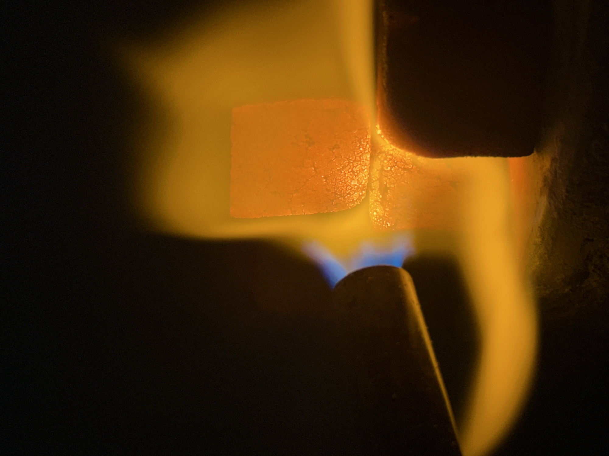 A close-up shot shows a sugar cube being melted by a blue flame from a lighter. The sugar cube is glowing orange. The image is dark with the exception of the flame and the glowing sugar cube.