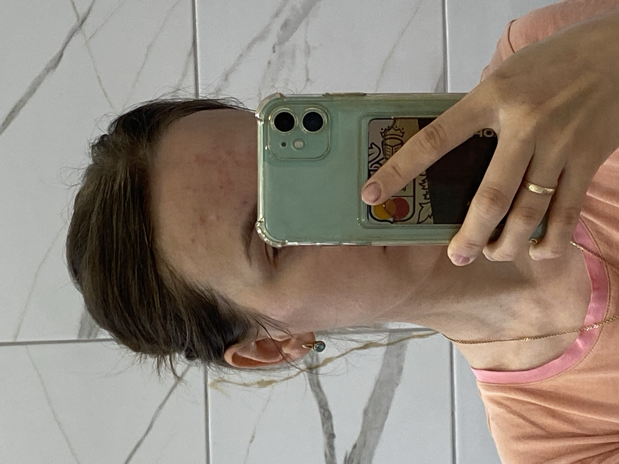 A young woman is taking a selfie in a bathroom. She has dark hair pulled back, a light complexion, and a few blemishes on her forehead. She is wearing a peach-colored t-shirt with a pink trim and a gold chain necklace. She is also wearing a gold ring on her left ring finger and a small gold earring with a teal stone in her left ear. She is holding a light green smartphone in her right hand, which is angled to capture her reflection. The background is a tiled wall with a marble pattern.
