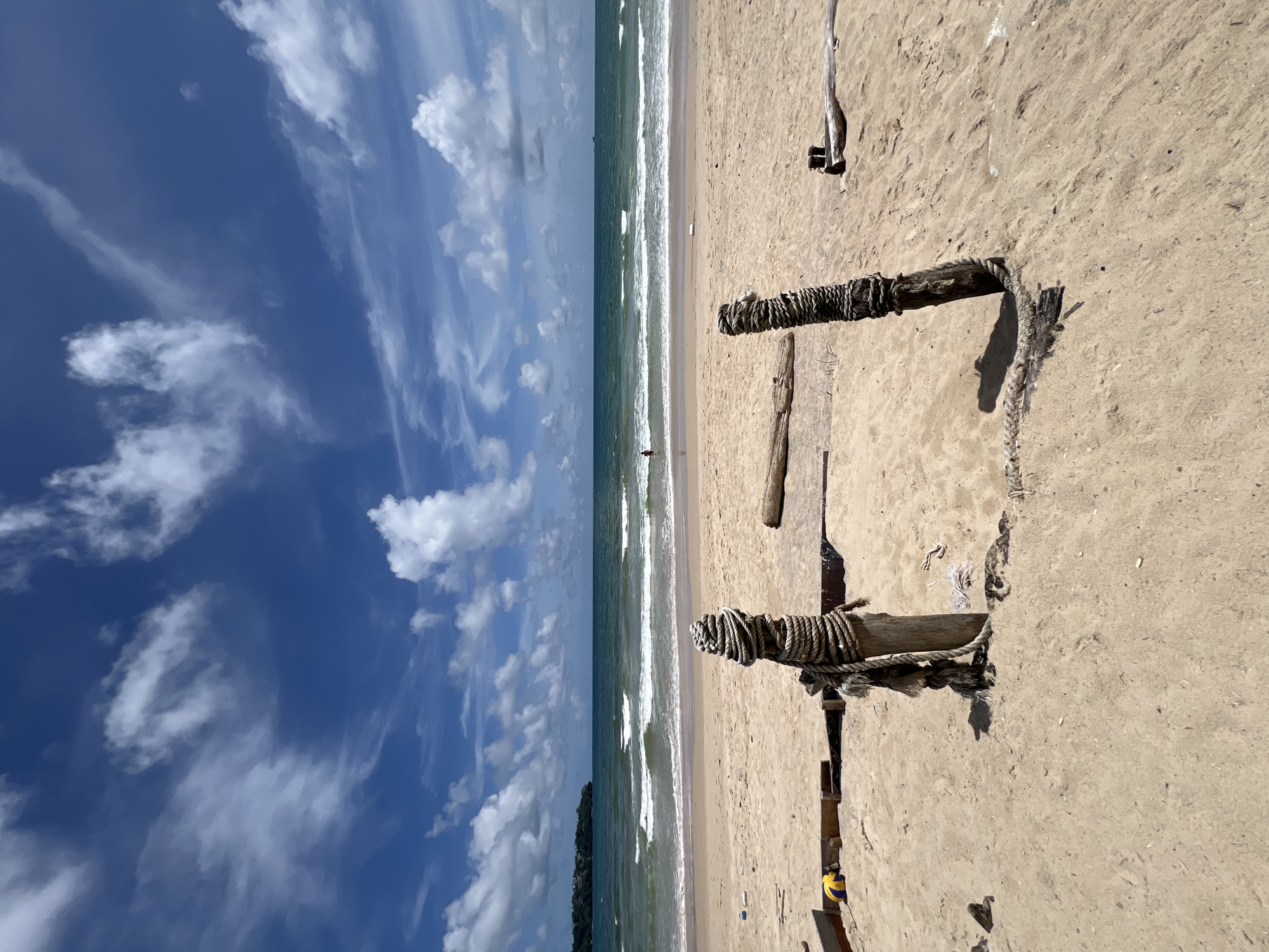 A wide shot of a sandy beach under a blue sky with white clouds. In the foreground, two weathered wooden posts are secured with thick rope. A piece of driftwood lies horizontally behind the posts. The sand is textured and marked with scattered debris. The middle ground features the ocean with gentle waves rolling onto the shore. A single person is visible in the water, standing near the breaking waves. The background shows a distant coastline with green foliage and a hint of structures. The overall scene conveys a tranquil, somewhat rustic beach setting.