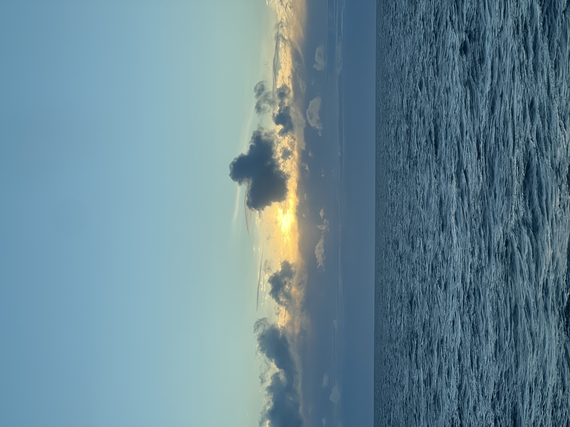 The image depicts a serene seascape at sunset. The sky transitions from a clear, light blue at the top to a mix of soft blues and yellows near the horizon, where the sun is partially obscured by clouds. Wispy clouds in shades of grey, white, and light yellow dot the sky, with a prominent dark grey cloud formation in the center catching the sun's golden rays. Below the sky, the vast expanse of the ocean stretches out, its surface covered in gentle ripples of deep blue. The horizon line is sharp and clear, dividing the sky and sea. There are no people, buildings, or specific landmarks visible, suggesting an open water location. The overall impression is one of natural beauty and tranquility.