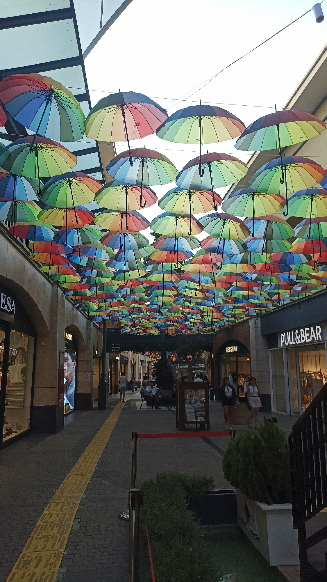 The image captures a vibrant outdoor shopping arcade or pedestrian street, characterized by an extensive canopy of colorful rainbow-striped umbrellas suspended overhead. These umbrellas, displaying hues of red, orange, yellow, green, and blue, create a cheerful and visually striking ceiling, diffusing the bright daylight from above.
The setting features commercial buildings on both sides, with arched passages and stone-clad facades giving an impression of a well-maintained, modern shopping district. On the left, shopfronts for "DESA" and "RAMSEY" are visible, while on the right, signs for "BERSHKA" and "PULL&BEAR" can be clearly seen.
Several people are scattered throughout the scene. Some are walking along the paved pathway, while others are seated on benches, enjoying the ambiance. A yellow tactile paving strip runs down the center of the walkway, indicating accessibility features. Planters with green foliage and decorative barriers with red ropes add to the street's landscaping.
The lighting suggests late afternoon or early evening, with a bright sky overhead creating soft shadows and a pleasant, diffused light beneath the umbrellas. The overall atmosphere appears relaxed and inviting, indicative of a leisure or shopping outing. The visible international brand names and the architectural style are consistent with urban shopping areas found in places like Yenişehir, Turkey.