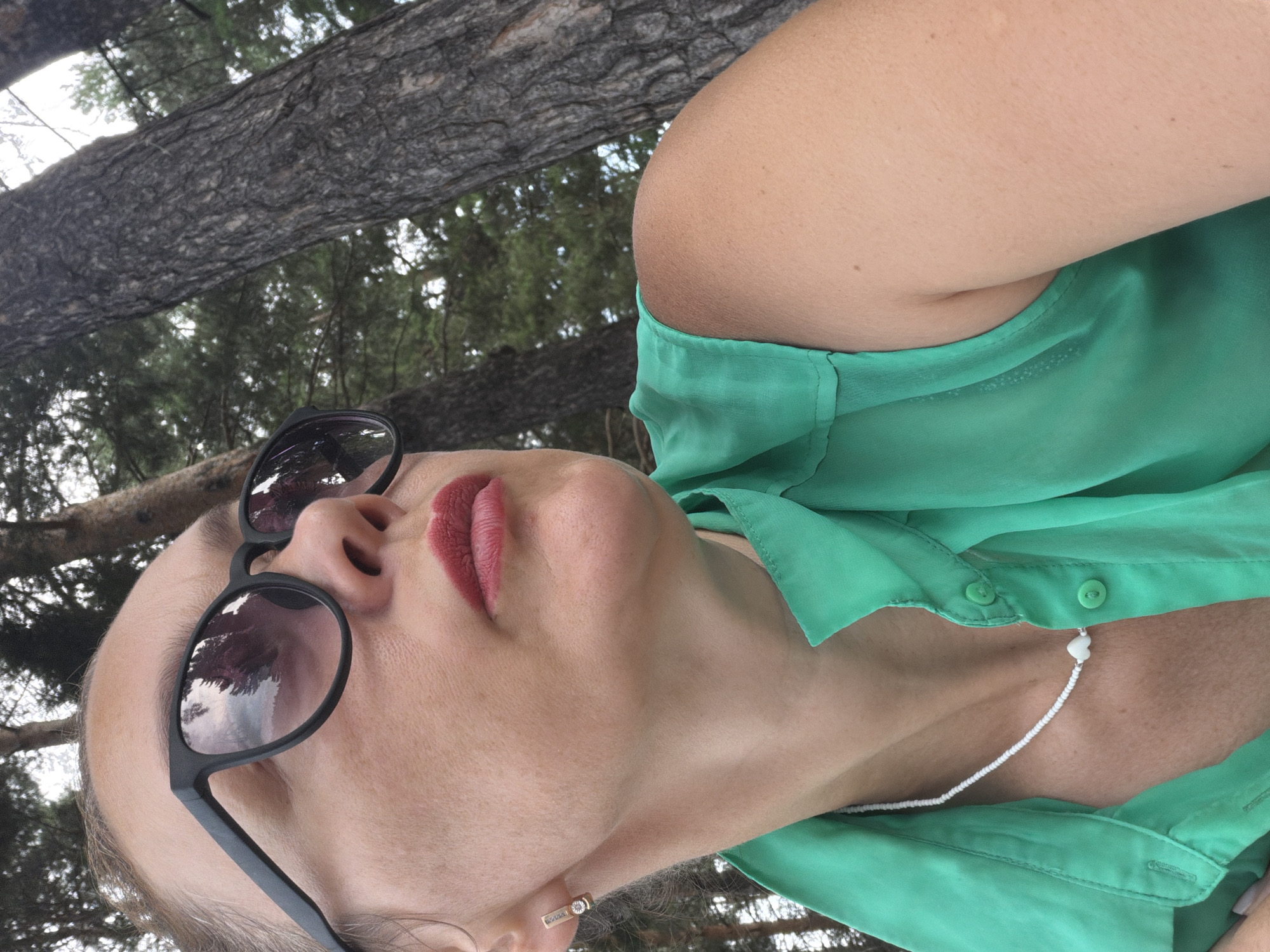 A woman with fair skin and dark hair pulled back is captured in a close-up, profile shot. She is wearing dark sunglasses with reflections of trees and a cloudy sky visible in the lenses. Her lips are painted with a matte red lipstick, and she is wearing a green sleeveless shirt with a collared neckline and two visible buttons. A delicate white beaded necklace with a white heart-shaped pendant rests on her collarbone. She has a small, rectangular gold earring with a small diamond in her left ear. The background consists of textured tree trunks and foliage, suggesting an outdoor, natural setting. The image appears to be taken in Chaikovsky, Russia, given the context of the query.