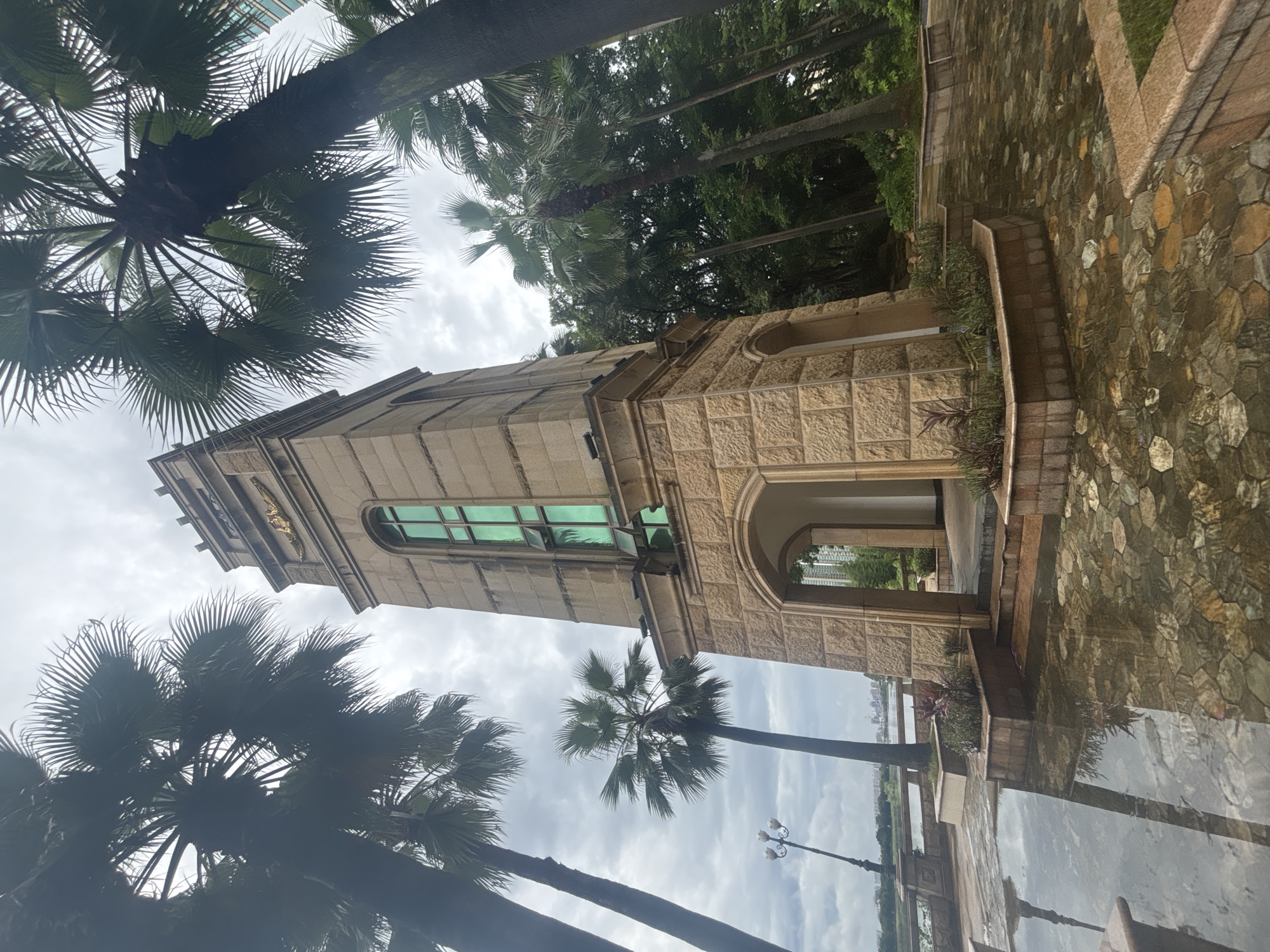 The image captures a tall, stone clock tower in Nanhai District, China, surrounded by lush palm trees. The tower, constructed from tan, square stone blocks, features a large arched entryway at its base, leading to a dark interior. Above the arch, a tall, green-paned window with open shutters is visible. At the very top of the tower, a golden emblem with wings can be seen against the overcast sky.
The ground in front of the tower is paved with hexagonal stones, and a shallow pool of water reflects the surrounding elements, including the clock tower and palm trees. Planters with small green plants are situated near the base of the tower. To the left of the tower, a lamppost with multiple lights stands near a stone railing overlooking a body of water. In the background, beyond the trees and railing, a cityscape with modern buildings is discernible. The overall atmosphere is serene, with the cloudy sky suggesting it might have recently rained. No people are present in the frame.