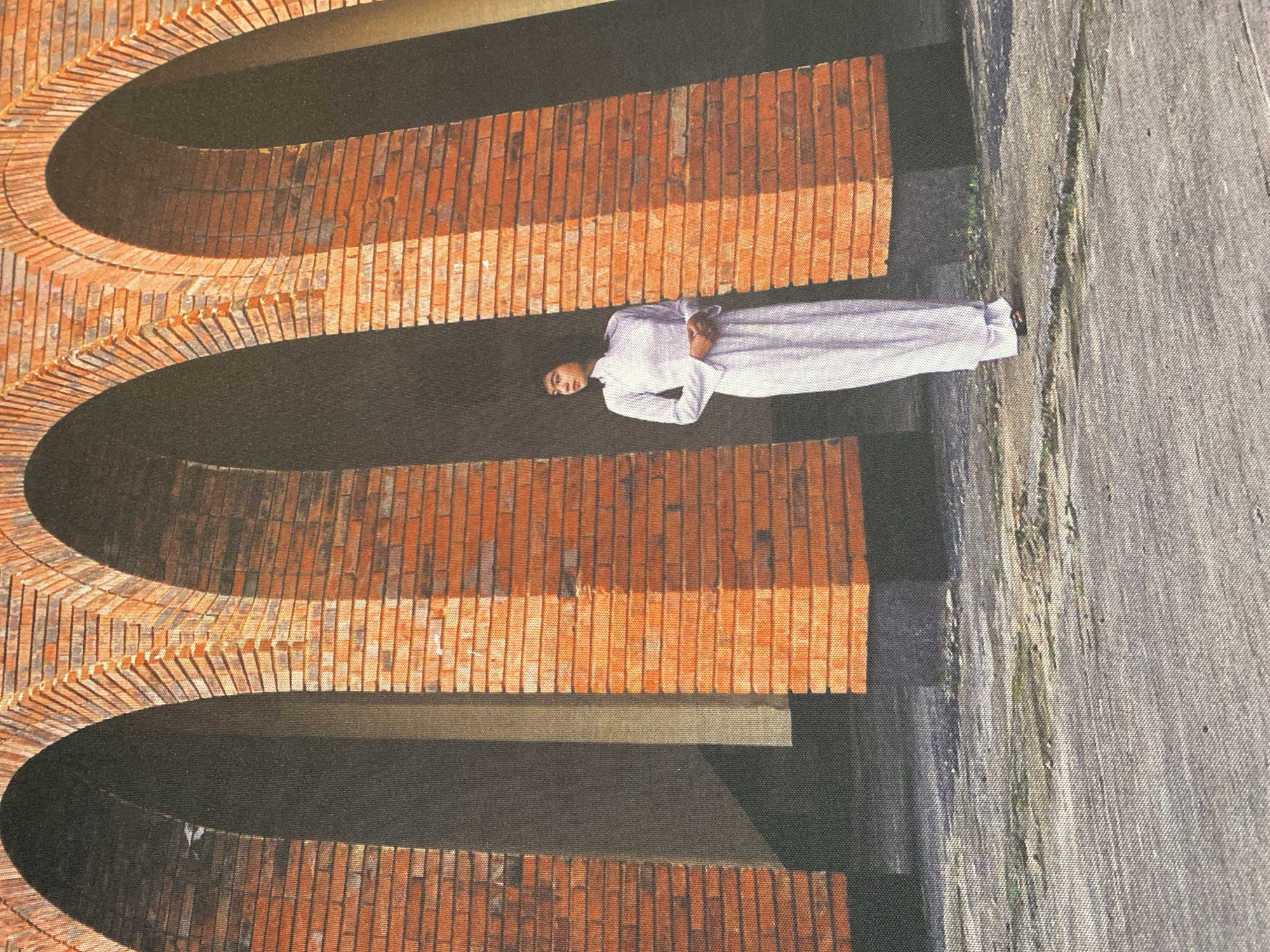 The image depicts a person in a white robe standing against a brick archway structure. The building is likely the Domain de Marie Convent in Da Lat, Vietnam, based on its architectural style. The structure consists of multiple stacked archways constructed of red bricks. The person is positioned against one of the archways, on a gray surface. The lighting suggests an outdoor daytime scene.