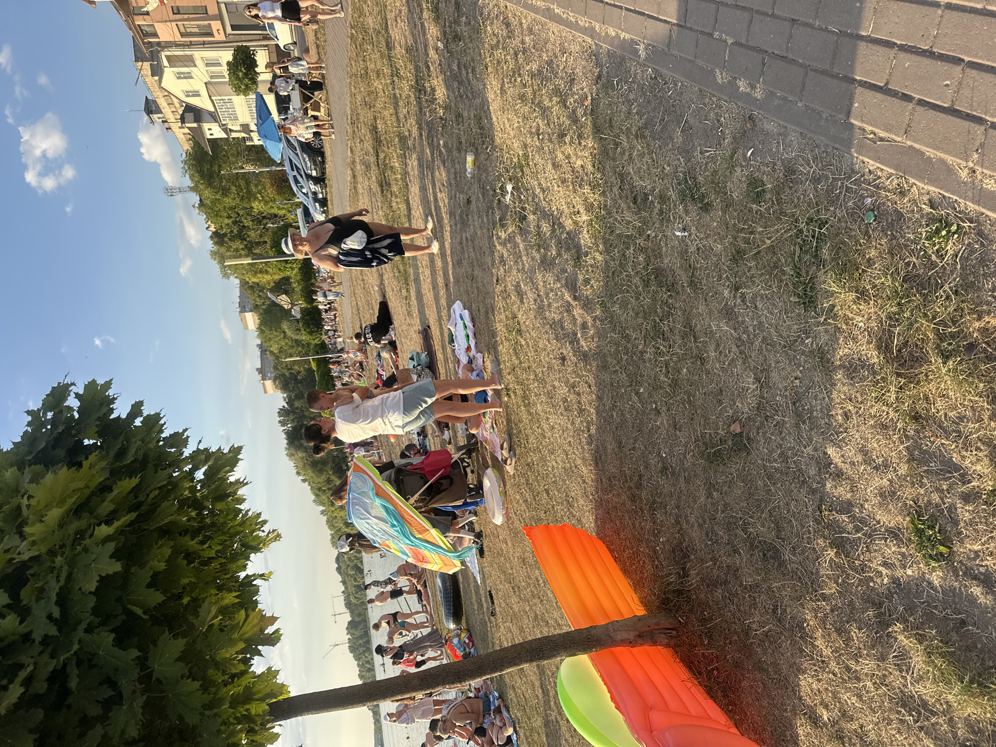 The image depicts a sunny day at a crowded public beach along the Ternopil Reservoir in Ternopil, Ukraine. The scene is characterized by a diverse group of people engaging in various leisure activities. In the foreground, a large, inflatable mattress, partially orange and partially green, lies on the dry, grassy ground near a tree trunk. Further back, the beach is populated with individuals and groups of varying ages, many of whom are dressed in swimwear.
Several people are seen relaxing on towels and blankets, some are standing, and others appear to be walking along the shoreline. A large, colorful beach umbrella is prominently displayed, suggesting a gathering or a family outing. Scattered throughout the scene are other beach accessories such as towels, bags, and smaller inflatable rings. The water of the reservoir is visible in the background, with more people enjoying the water and the shoreline.
The environment consists of a strip of dry grass and sand leading to the water. Beyond the beach, a city skyline is visible, with buildings and trees lining the elevated bank. Cars are parked along a paved area adjacent to the beach, indicating vehicular access to the location. The sky is clear blue with a few scattered clouds, and the lighting suggests it is late afternoon or early evening, with the sun casting long shadows. The overall impression is of a lively, recreational public space during a warm weather season.