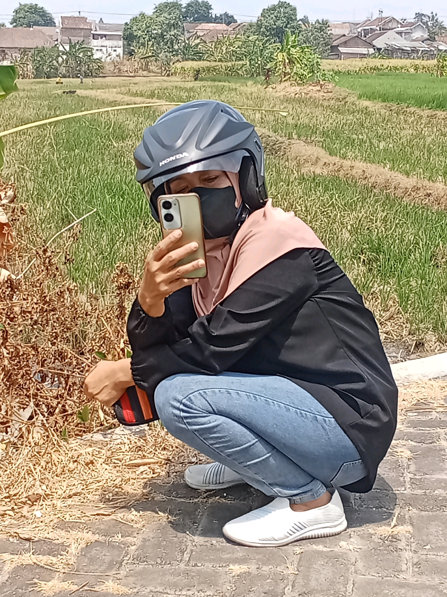 A person wearing a black helmet, black face mask, light pink hijab, black jacket, and blue jeans is squatting on a brick path, taking a selfie with a gold smartphone. The person is positioned in the foreground, with their body angled towards the left. In the background, there is a field of green grass and some houses. The scene appears to be in Sukowinangun, Indonesia, given the rice paddies and tropical vegetation. The lighting suggests it is daytime, with bright sunlight. The overall atmosphere is casual and outdoor.