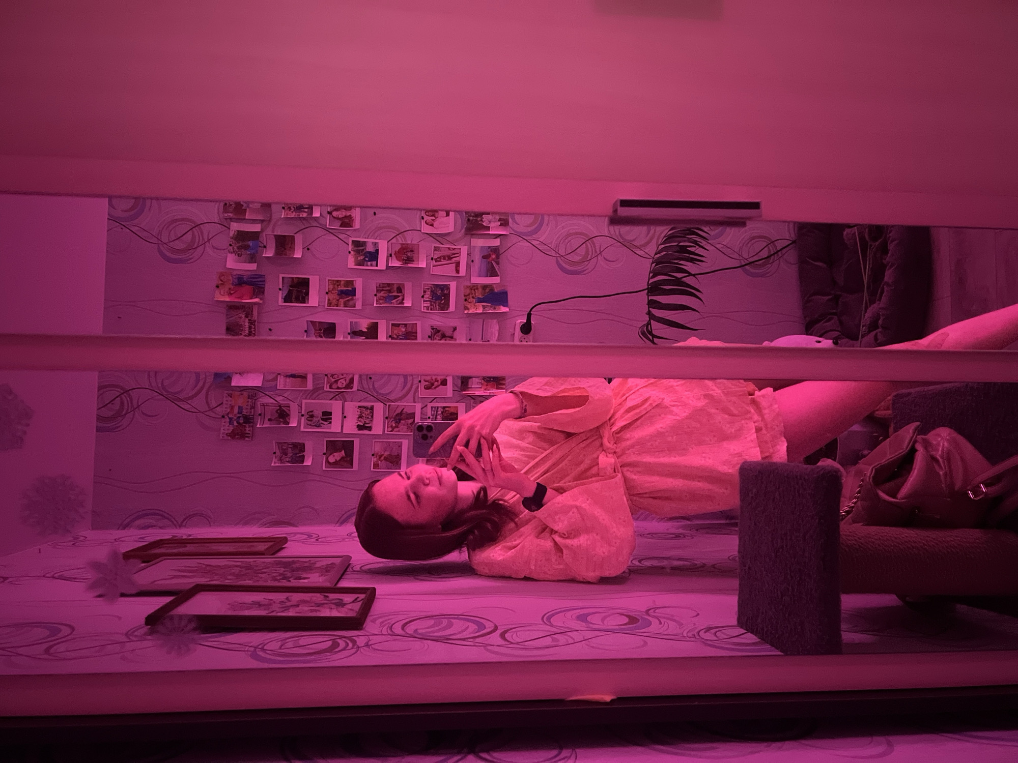 A person lies on a bed in a room illuminated with pink light. The location is likely Chaikovsky, Russia, but this cannot be definitively determined from the image. The room's wallpaper features a pattern of waves and is decorated with pinned-up photographs. Additional objects in the room include picture frames, a chair, and a purse. The person is holding a smartphone and appears to be looking at it.