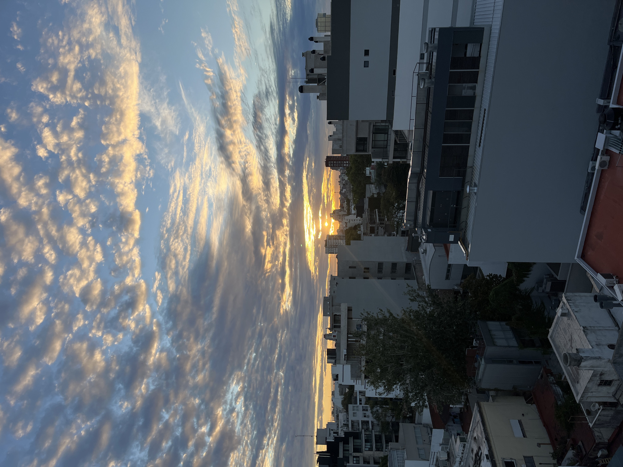 The image depicts a sunset over a city. The sky is filled with streaks of clouds illuminated by the setting sun. The city consists of closely packed buildings of varying heights, appearing to be an urban residential area. The location is Buenos Aires, Argentina. The image is taken from an elevated point of view, looking down onto the city. There are no people visible.