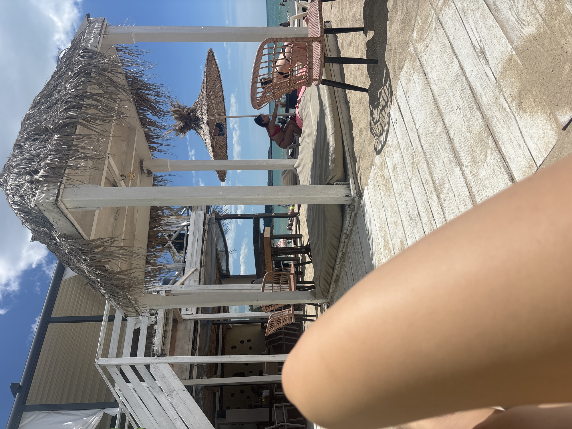 This is a first-person perspective photograph taken on a sunny day at a beachside establishment in Varna, Bulgaria. The foreground is dominated by the photographer's leg, positioned to the left and partially obscuring the wooden deck flooring. The deck, made of weathered white planks, is partially covered with sand. In the midground, a thatched-roof cabana stands prominently, supported by white pillars. Beneath the cabana, there are large cushions or mattresses laid out on the sand. To the right of the cabana, a pink rattan chair with black legs is visible, casting a patterned shadow on the deck. In the background, two women are seen under a straw umbrella; one, wearing a red bikini, is reaching towards a table, and the other, in a black bikini, is also interacting with the table. Further back, there are more beach umbrellas, beachgoers, and the sea. To the left of the cabana, a two-story wooden structure with white stairs and railings is partially visible, suggesting a building or a raised platform. The sky is blue with scattered white clouds.