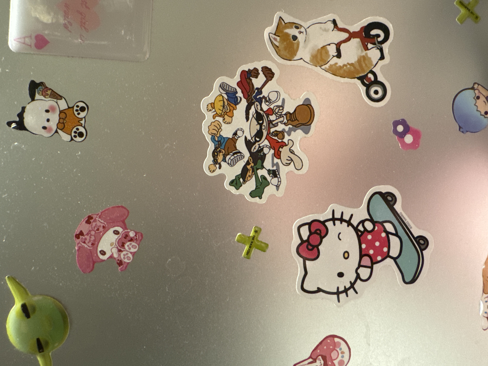 In the city of Бараўлянскі сельскі Савет, Belarus, a metallic surface, likely a refrigerator door, is adorned with various cartoon stickers. On the top left, a green character with large ears and black eyes is depicted. To its right, a pink character with a bow and bell is positioned. Further right, a white dog-like character in an orange outfit holds an ice cream cone. A partially visible playing card with an "A" and a heart is to the right of the dog. Below the green character, a green cross-shaped sticker with Cyrillic text is visible. In the center of the image, a group of animated characters in action poses are clustered together. To the right of this group, a cat sticker shows a cat riding a tricycle. Below the cat sticker, a small pink flower sticker with purple accents is present. A large sticker of Hello Kitty riding a skateboard is situated in the lower left quadrant of the image. A blue-haired character is at the bottom center, and a yellow cross is at the bottom right.
