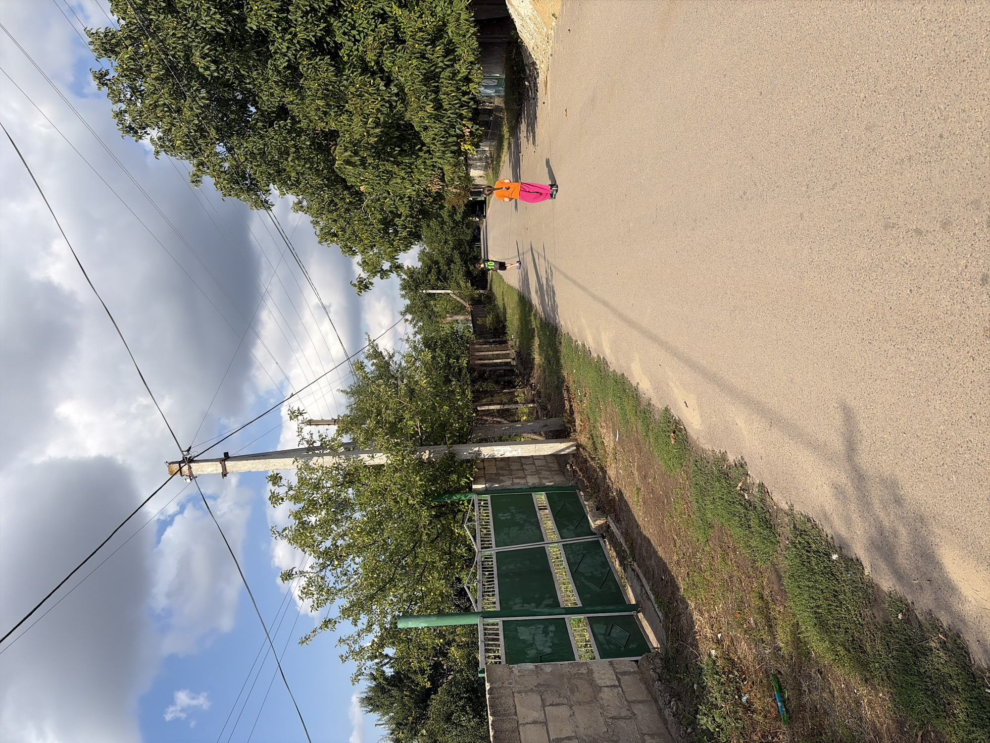 A rural road in Oxentea, Moldova, is captured on a partly cloudy day. The road is paved and stretches into the distance. On the left side of the road, there is a green metal gate set within a rough stone wall, bordered by overgrown grass and vegetation. A concrete utility pole with overhead wires stands near the gate. On the right side of the road, mature trees with dense green foliage line the path, casting shadows. In the middle of the road, two children are present. One child, wearing a black t-shirt with the number "10" and black shorts, is walking away from the camera. Another child, wearing an orange t-shirt and bright pink pants, is riding a scooter away from the camera. The background shows more trees and a few distant buildings, suggesting a village or rural setting.