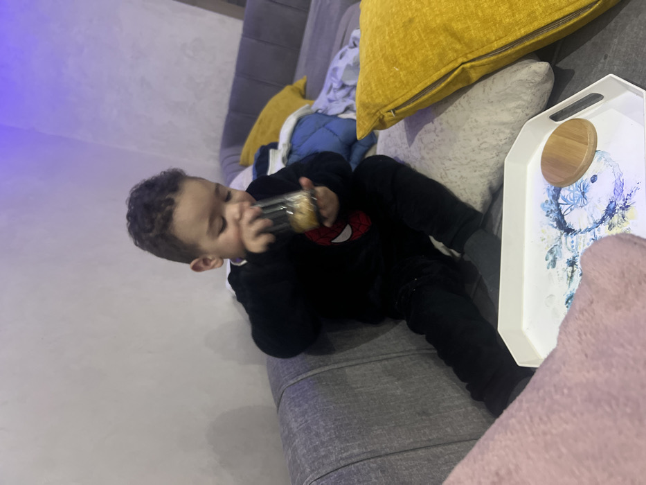 A toddler wearing a black Spider-Man sweatshirt and black pants reclines on a grey couch, drinking from a bottle. He is surrounded by throw pillows, including yellow and grey ones. A white tray rests beside him, decorated with a blue and white floral design and topped with a round, wooden object. The background consists of a plain, light-colored wall and a portion of the grey couch.