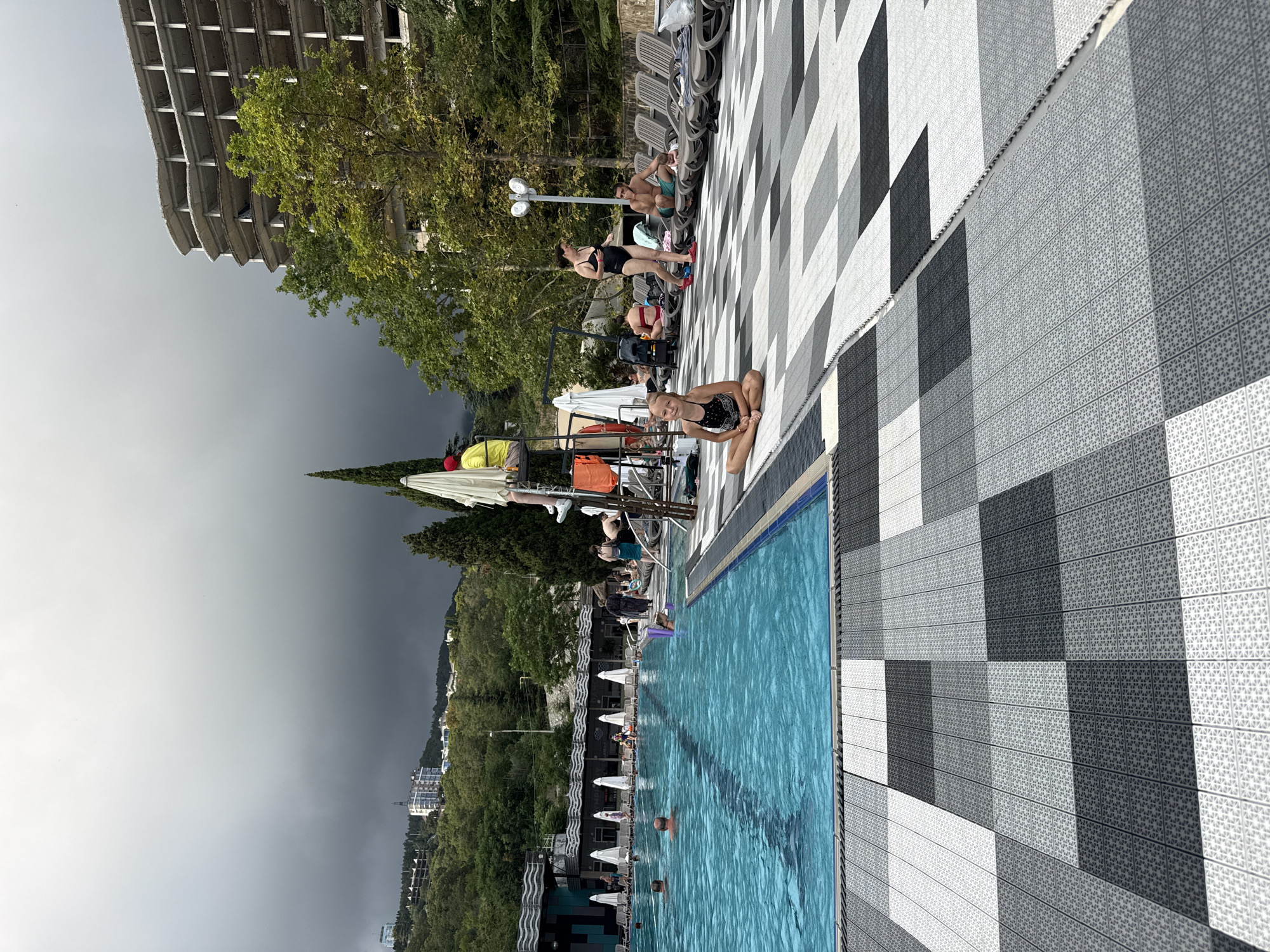 The image depicts an outdoor swimming pool area in Yalta, Ukraine, on a cloudy day. A young girl in a black and white swimsuit sits cross-legged on the patterned tiled ground in the foreground. The pool is filled with blue water and has lane markers. Several people are in and around the pool, including men swimming and people relaxing on lounge chairs. A lifeguard in a yellow shirt and red hat is seated on a raised platform. In the background, lush green trees and hills are visible, with a partially constructed, multi-story building dominating the skyline. The sky is overcast with dark, heavy clouds.