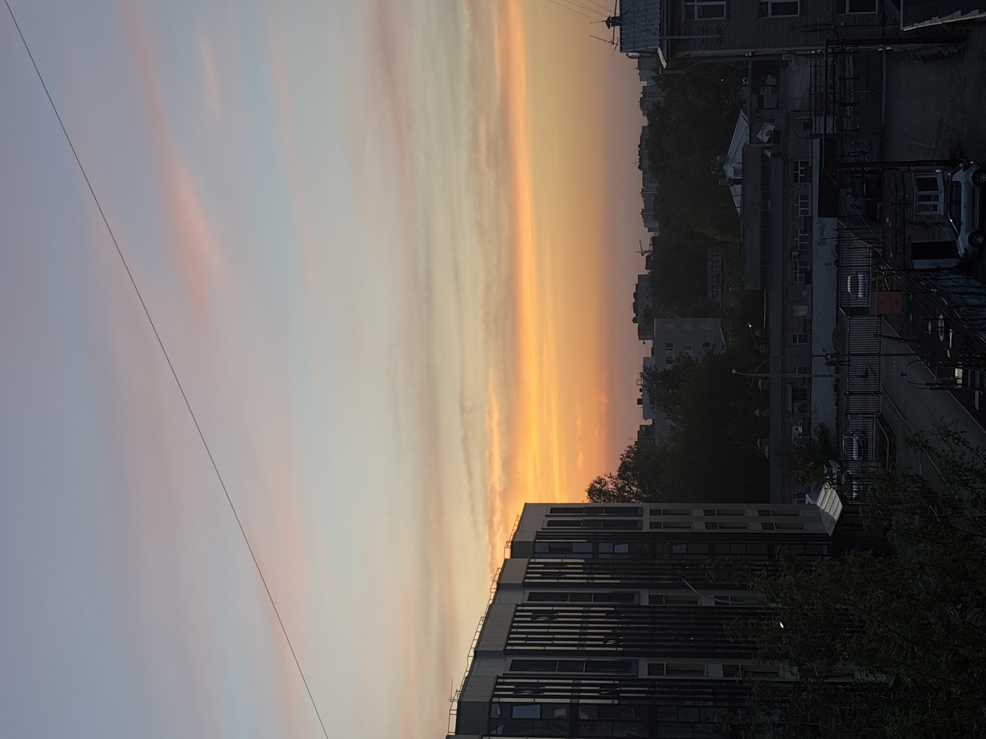A vertical shot reveals a tranquil cityscape in Perm, Russia, bathed in the warm hues of a sunset. The sky showcases a gradient of colors, transitioning from a pale lavender to a vibrant orange near the horizon. Light, wispy clouds stretch across the sky, adding texture to the serene expanse.
The cityscape is a mix of residential buildings and urban infrastructure. Modern apartment buildings with rows of windows are visible in the foreground. In the middle ground, dense greenery dots the landscape, with trees interspersed among older, more traditional buildings. The streets are narrow and lined with parked cars, suggesting a quiet evening in the city. A power line cuts diagonally across the sky.