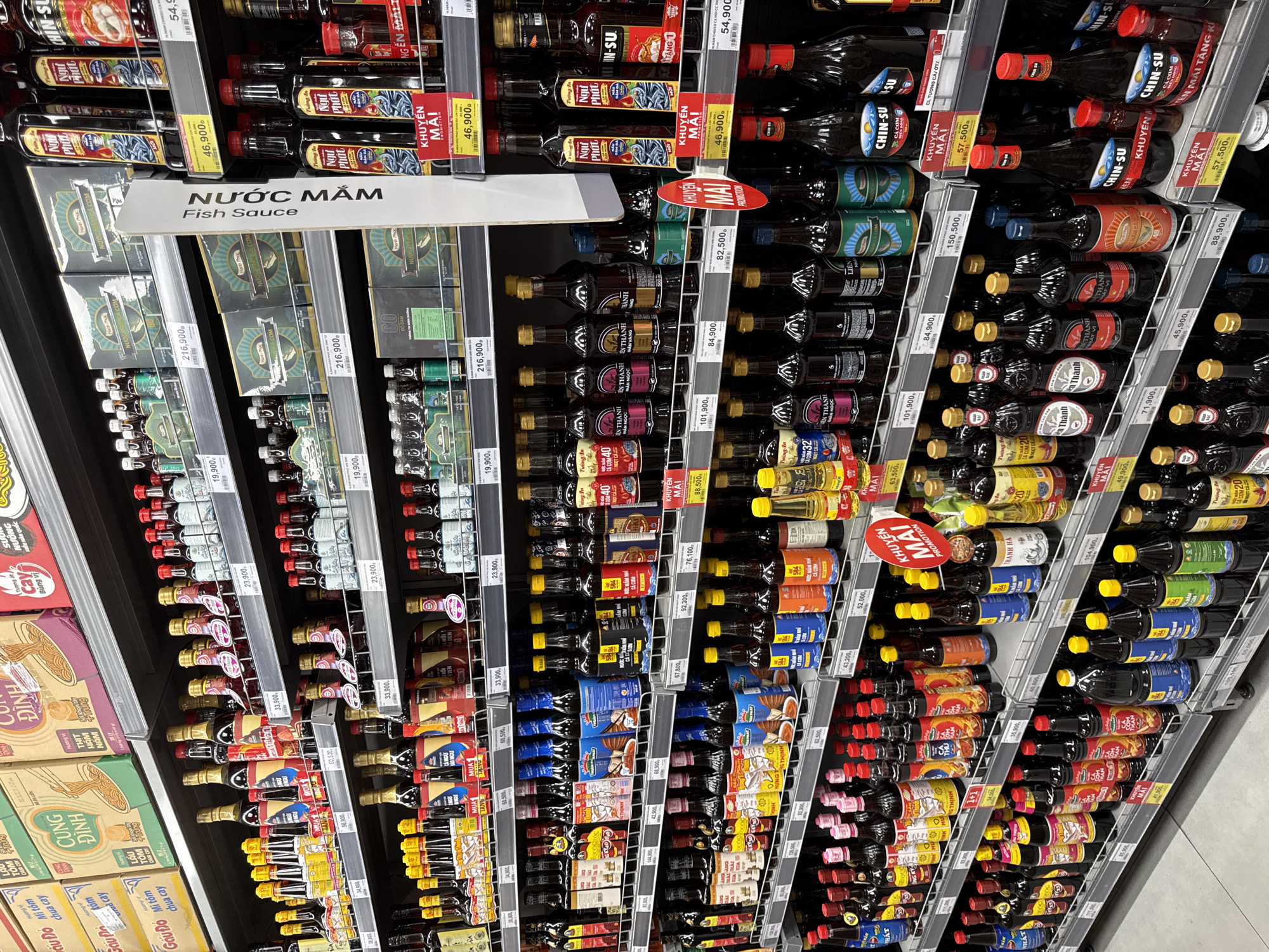 The image shows a brightly lit grocery store aisle, specifically focusing on shelves stocked with various bottles of fish sauce and other condiments. The products are neatly arranged in rows, with many featuring Vietnamese labels. The shelves are made of metal, and the store appears to be a modern supermarket with clean displays and price tags visible. The overall impression is one of an organized and well-stocked retail environment. There are no people or specific landmarks visible that would indicate a precise location within Nha Trang, Vietnam.