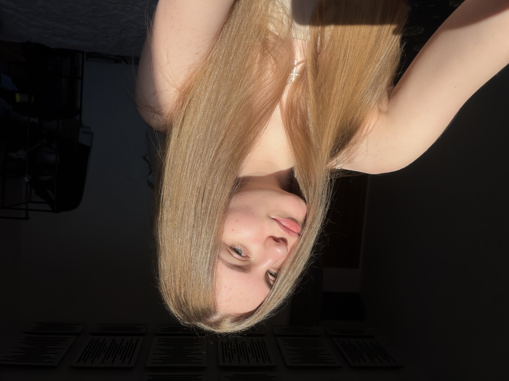 A young woman with long, blonde hair is depicted in an indoor setting, possibly her room. The image is oriented sideways, with the woman's head pointing downwards. She is wearing a light-colored top, and a sparkling necklace is visible around her neck. Her gaze is directed towards the camera, and her lips are slightly parted. The lighting suggests it is daytime, with sunlight casting shadows in the background. In the foreground, a set of items with horizontal lines are laid out. The overall mood is relaxed and casual. The location is specified as Dnipro, Ukraine.