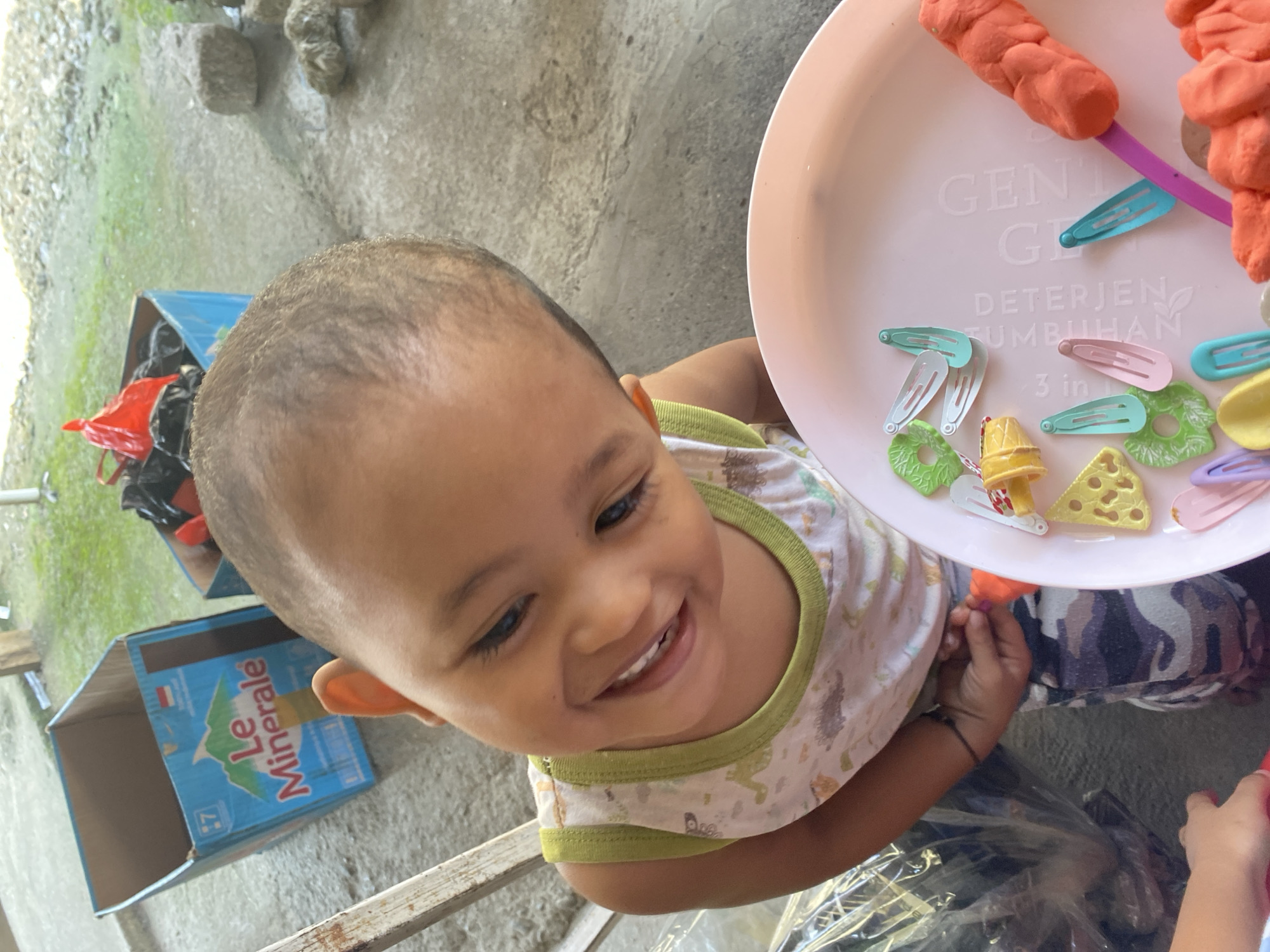 A child with dark hair and brown eyes is looking down and smiling at a pink tray of objects. The child is wearing a white tank top with a green trim and a pattern of dinosaurs. The tray contains several small colorful hair clips and some pieces of orange playdough. The background shows a concrete floor and some cardboard boxes. One box has the "Le Minerale" logo on it. The setting appears to be an outdoor area in Ta', Indonesia.