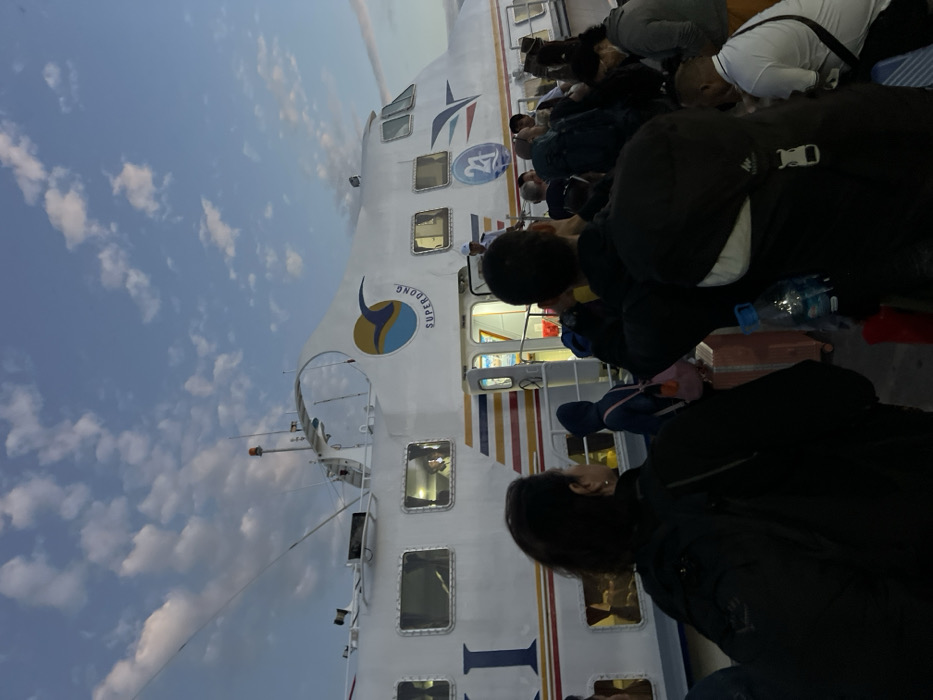 The image depicts a crowded deck scene aboard a white passenger ferry, viewed from a low-angle perspective looking forward. Numerous passengers, mostly adults, are visible in the foreground and midground. Many are seated or lying down on the deck, often with their backs to the camera or heads bowed, suggesting they are resting, sleeping, or simply waiting during the journey. Various pieces of luggage, including backpacks and suitcases, are scattered around them.

The ferry itself is a prominent subject, characterized by its white hull, multiple rectangular windows with illuminated interiors, and a horizontal stripe of red, yellow, blue, and white along its lower side. A clear circular logo on the ferry's side prominently displays the text "SUPERDONG," identifying it as part of a known Vietnamese ferry operator. Another stylized star logo is also visible further along the vessel.

The upper portion of the image reveals a clear, light blue sky with scattered white cumulus clouds, indicating fair weather conditions. The lighting, combining soft natural light from the sky with artificial lights visible inside the ferry, suggests the time of day is either early morning (dawn) or late afternoon/early evening (dusk). The overall atmosphere appears calm, reflecting the typical experience of travelers undertaking a long or overnight ferry journey. Given the "SUPERDONG" branding and the provided location context of Hà Tiên, Vietnam, the ferry is likely operating routes in the Gulf of Thailand, possibly to or from Phú Quốc Island.