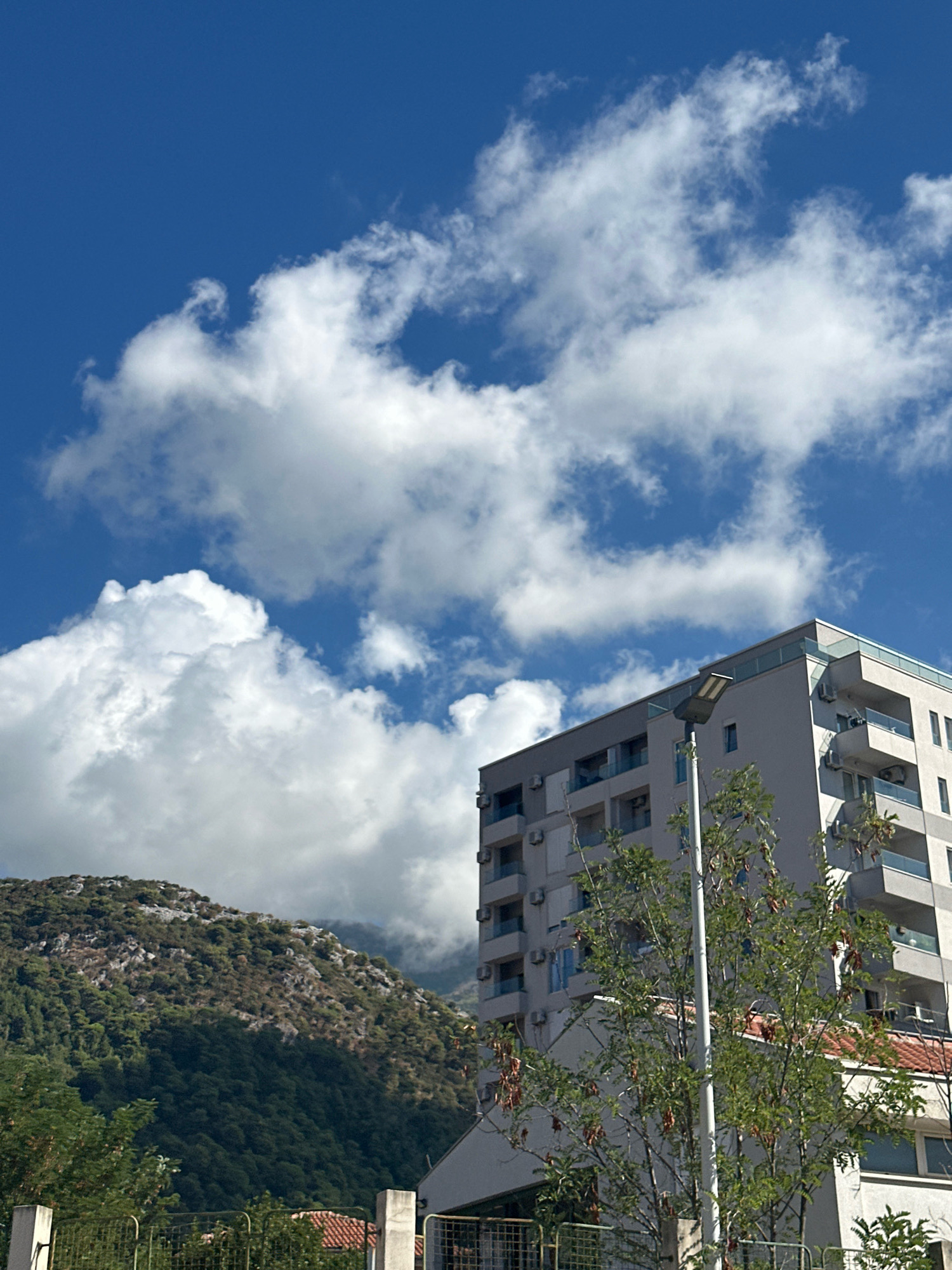 In Budva, Montenegro, a modern apartment building stands against a backdrop of lush, green mountains under a vibrant blue sky filled with dynamic white clouds. The building, with its light-colored facade, features multiple balconies and air conditioning units. A tall lamppost is positioned in front of the building, partially obscuring a leafy tree with seed pods. The mountainous terrain is densely covered with dark green trees, with patches of lighter rock visible on its slopes. In the foreground, a metal fence runs horizontally, with hints of red-tiled roofs visible behind it. The scene captures a clear, sunny day with dramatic cloud formations adding depth to the sky.