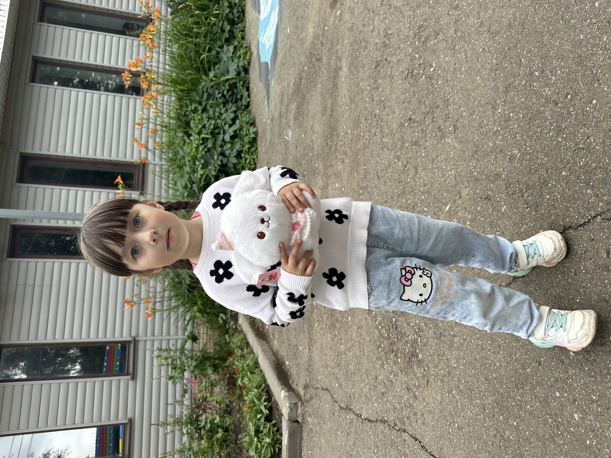 This is a vertical, eye-level, full-body photograph of a young girl standing outdoors. She is holding a white, fluffy stuffed animal with a pink bow in front of her chest. The girl has brown hair styled in pigtails and is wearing a white sweater with black floral patterns and light blue jeans. Her jeans are adorned with a large Hello Kitty graphic on the right leg and some light orange lettering on the left. She is also wearing white sneakers with multicolored soles.
The girl is standing on an asphalt surface, with a gray concrete curb to her left. Behind her is a building with white siding and horizontal lines, featuring several rectangular windows. Orange flowers and green foliage are visible alongside the building. The lighting suggests it is daytime, and the weather appears to be overcast, given the soft shadows. The location context is Chaikovsky, Russia. The girl's expression is neutral.