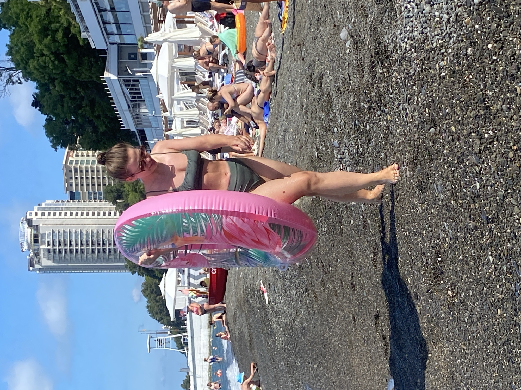 A young woman in a dark green bikini walks on a pebble beach in Sochi, Russia, holding a large inflatable pink ring with a flamingo design. She is wearing sunglasses and her hair is pulled up in a bun. The beach is crowded with people sunbathing and swimming. In the background, there are hotels, trees, and the Black Sea. The sky is clear and blue. The overall impression is of a sunny day at a popular tourist destination.