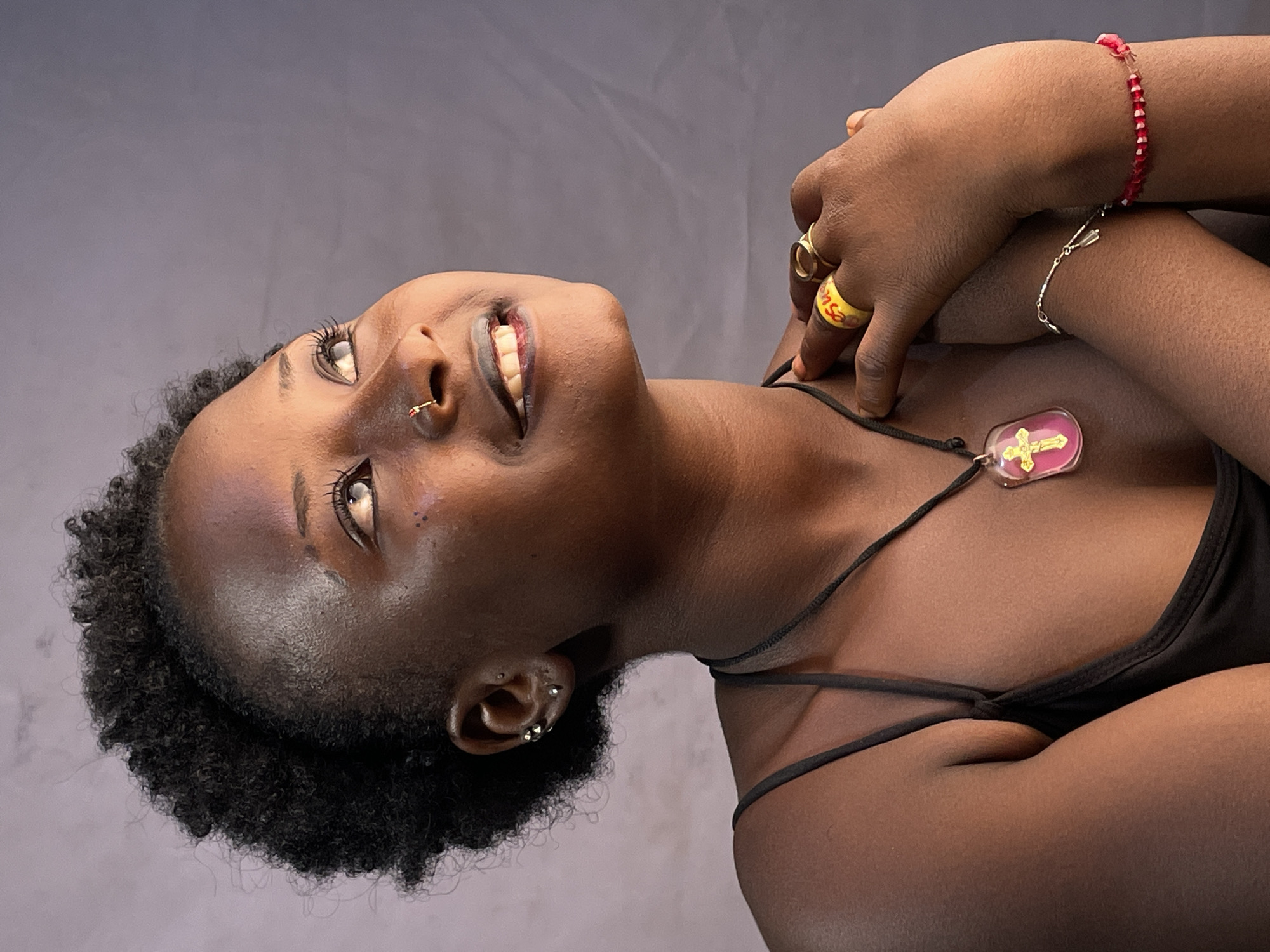 A close-up studio shot shows a dark-skinned woman angled upwards, smiling and looking off-camera. Her hair is short, dark, and curly. She has multiple piercings in her ears and a nose ring. She wears a black tank top and a necklace with a pendant featuring a cross on a pink background. On her hands, she has several rings and bracelets, including a yellow ring with writing, a silver chain bracelet, and a red beaded bracelet. The background is a smooth, neutral grey. The location and city of Jos, Nigeria cannot be confirmed from the given image.