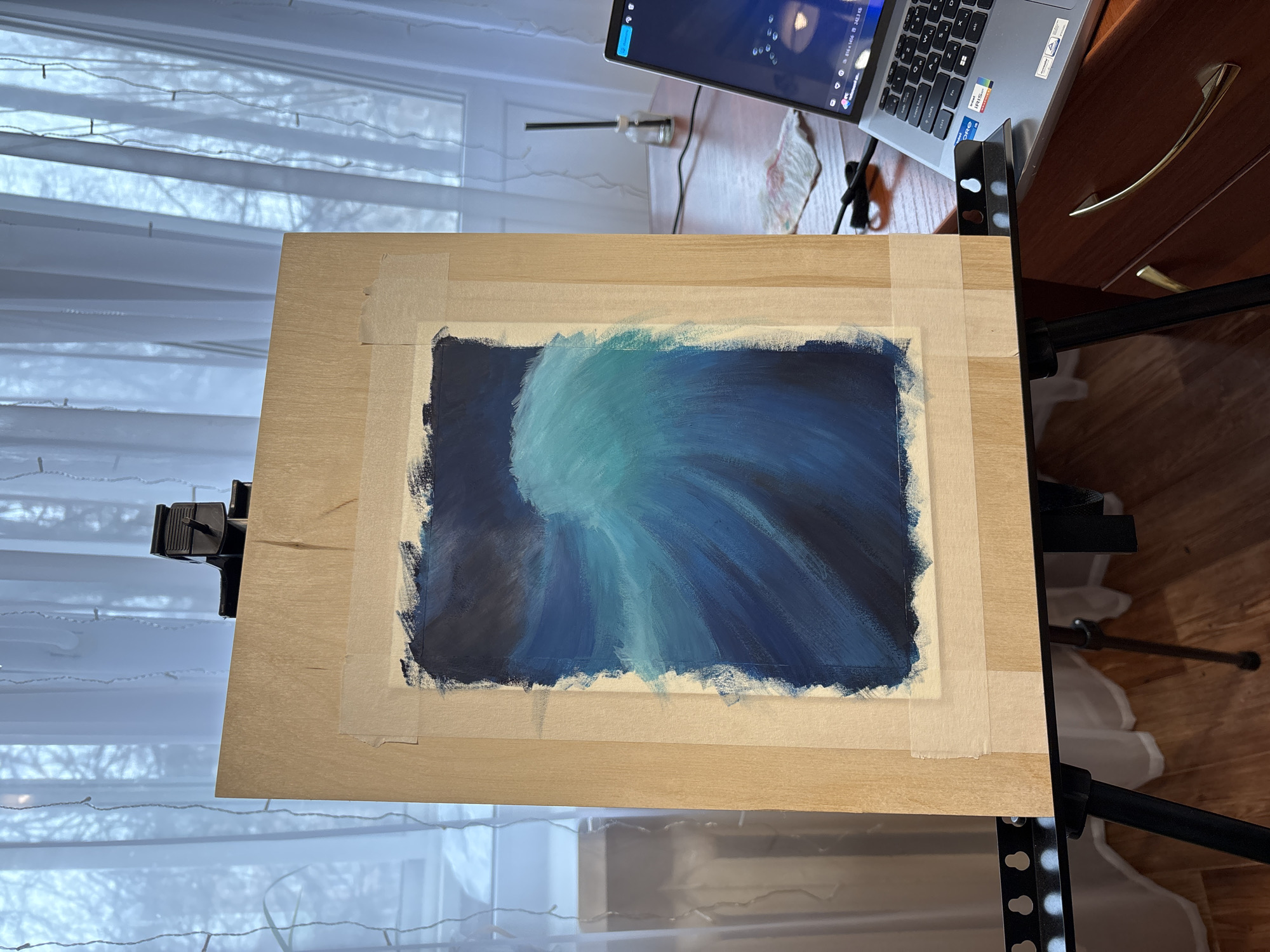 A photograph shows an art canvas mounted on a black easel. The canvas displays a painting with blue hues, seemingly an abstract composition. The canvas is bordered with beige masking tape and is attached to a wooden board. Positioned behind the canvas is a desk with a laptop, featuring a video playing on its screen. Adjacent to the desk is a wooden cabinet with golden handles. In the background, sheer white curtains hang over a window, allowing diffused natural light into the room. Based on the setting, I cannot verify the location of this image as the city of Izhevsk, Russia.