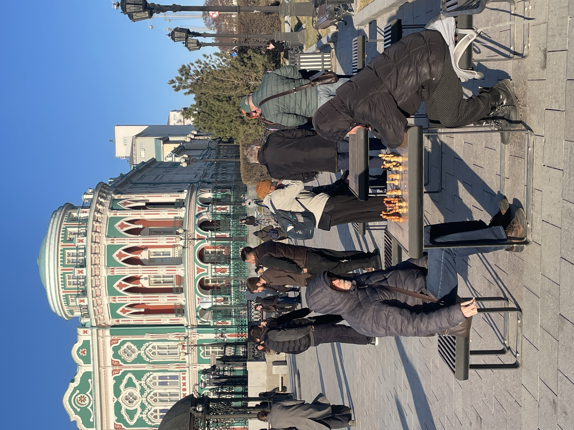 The image presents a lively outdoor scene in Yekaterinburg, Russia, under a clear blue sky. The focal point is a gathering of people engaged in and observing chess games in a public square or park.
In the foreground and mid-ground, multiple individuals are engrossed in playing chess at dedicated outdoor tables. Players, dressed in heavy winter jackets, puffer coats, and hats, lean over the boards, concentrating on their moves with wooden chess pieces. Spectators stand closely around the tables, watching the games. Other people are seen walking casually along the paved area, which features gray rectangular tiles.
To the left, a striking, multi-story building dominates the architectural landscape. It features distinctive green, white, and red facades with elaborate architectural details, including arched windows and decorative moldings, capped by a large, light-colored dome. The building's design suggests a significant landmark.
The scene is set in an urban environment, characterized by ornate black lampposts, public benches, and scattered trees, some coniferous and others deciduous. Bright sunlight casts long shadows, indicating either morning or late afternoon. Despite the clear sky, the warm clothing worn by most people suggests a cool or cold day. The overall atmosphere is one of focused concentration on the chess games, interspersed with casual pedestrian activity. No discernible text is visible in the image.