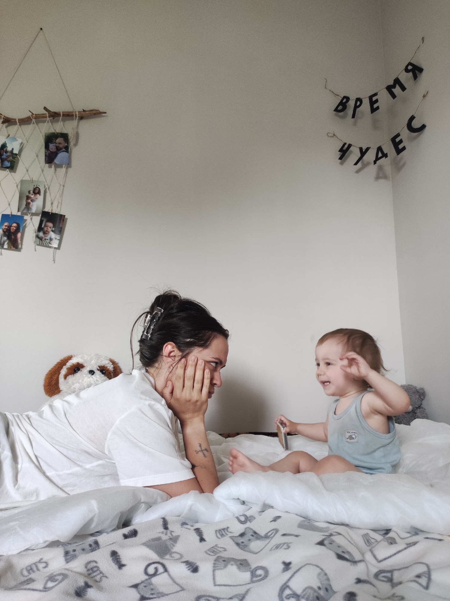 A young woman and a toddler are on a bed covered with a patterned blanket featuring drawings of cats and fish. The woman, with dark hair pulled back with a clip, is lying on her side, propping her head up with her hand and looking towards the toddler. The toddler is sitting up, wearing a light blue tank top, and holding a small object. A stuffed animal is behind the woman. The wall has a handmade photo display with a branch and netting, and a banner hanging on the upper right.