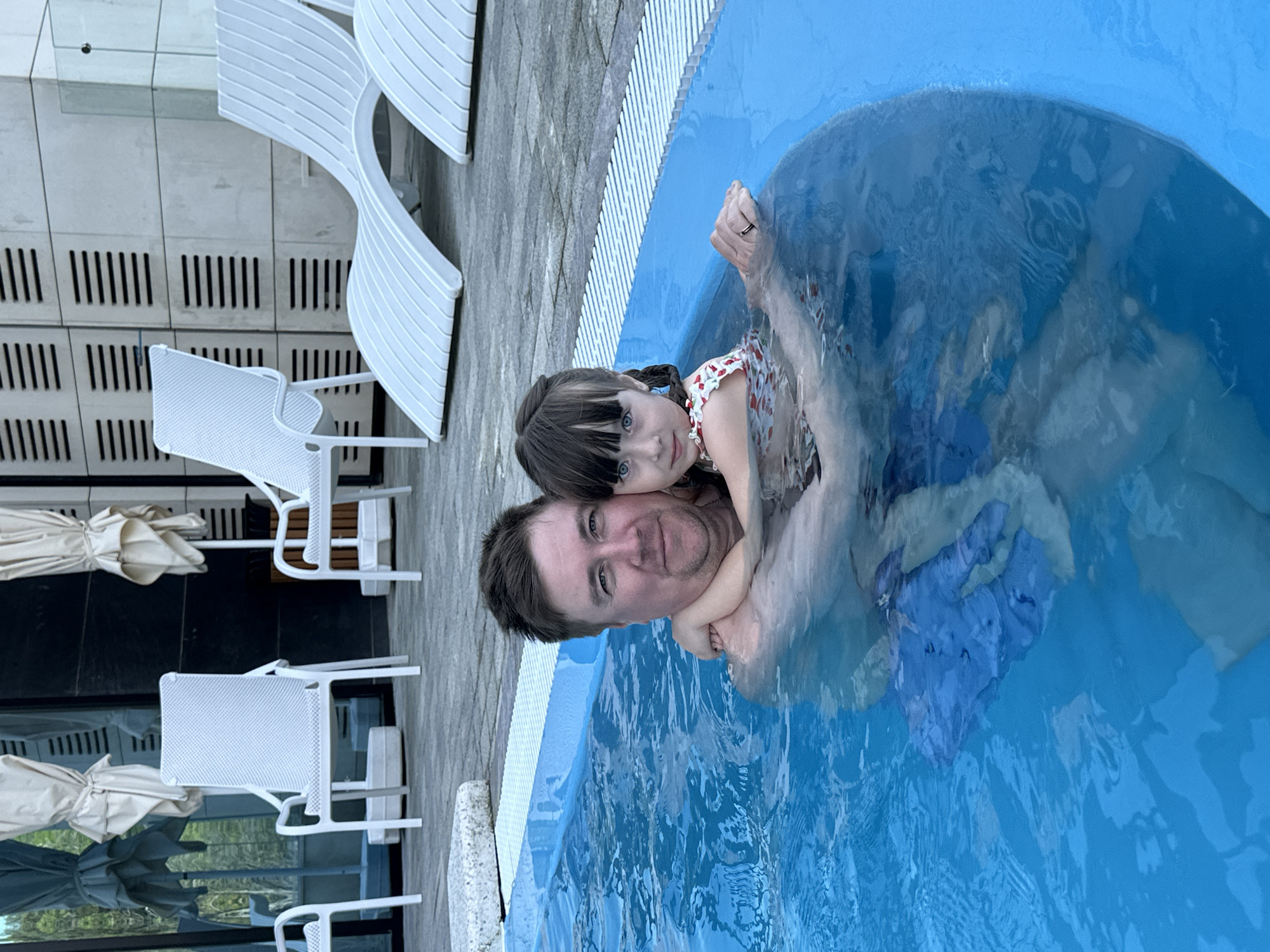 A Caucasian male is in a swimming pool holding a young Caucasian female. The man has short brown hair, blue eyes, and a light complexion. The girl has long brown hair in braids, blue eyes, and a light complexion. The water is blue and there is a blue cloth in the water as well. There are white chairs and umbrellas in the background. The location is Sochi, Russia.