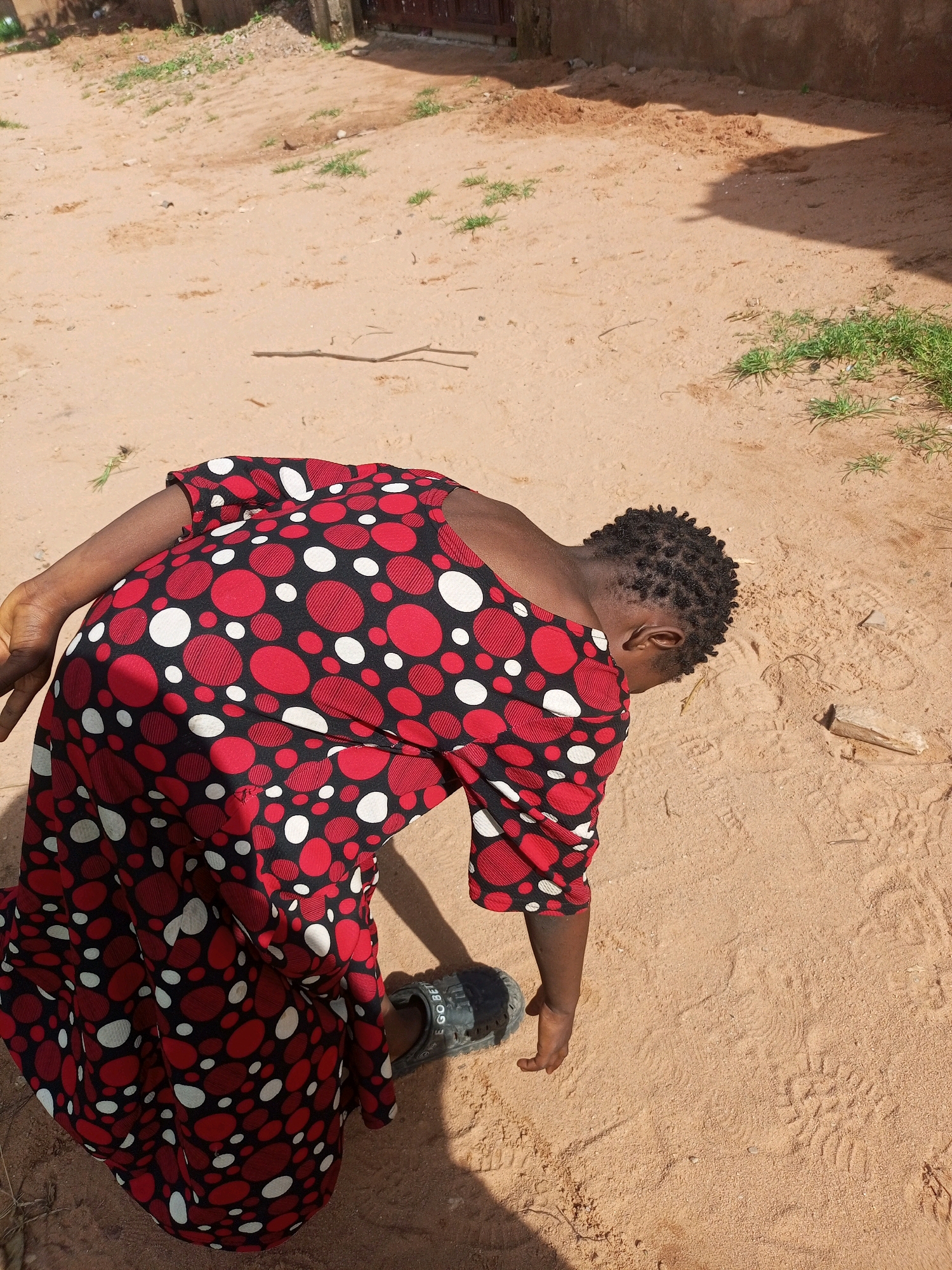 A person wearing a red and white polka dot dress is bent over in a sandy area. The person's back is to the camera, and their head is down. There is a single black sandal on the ground near the person's feet. The environment appears to be an outdoor setting with sand, dirt, and some sparse vegetation. The lighting suggests it is daytime.