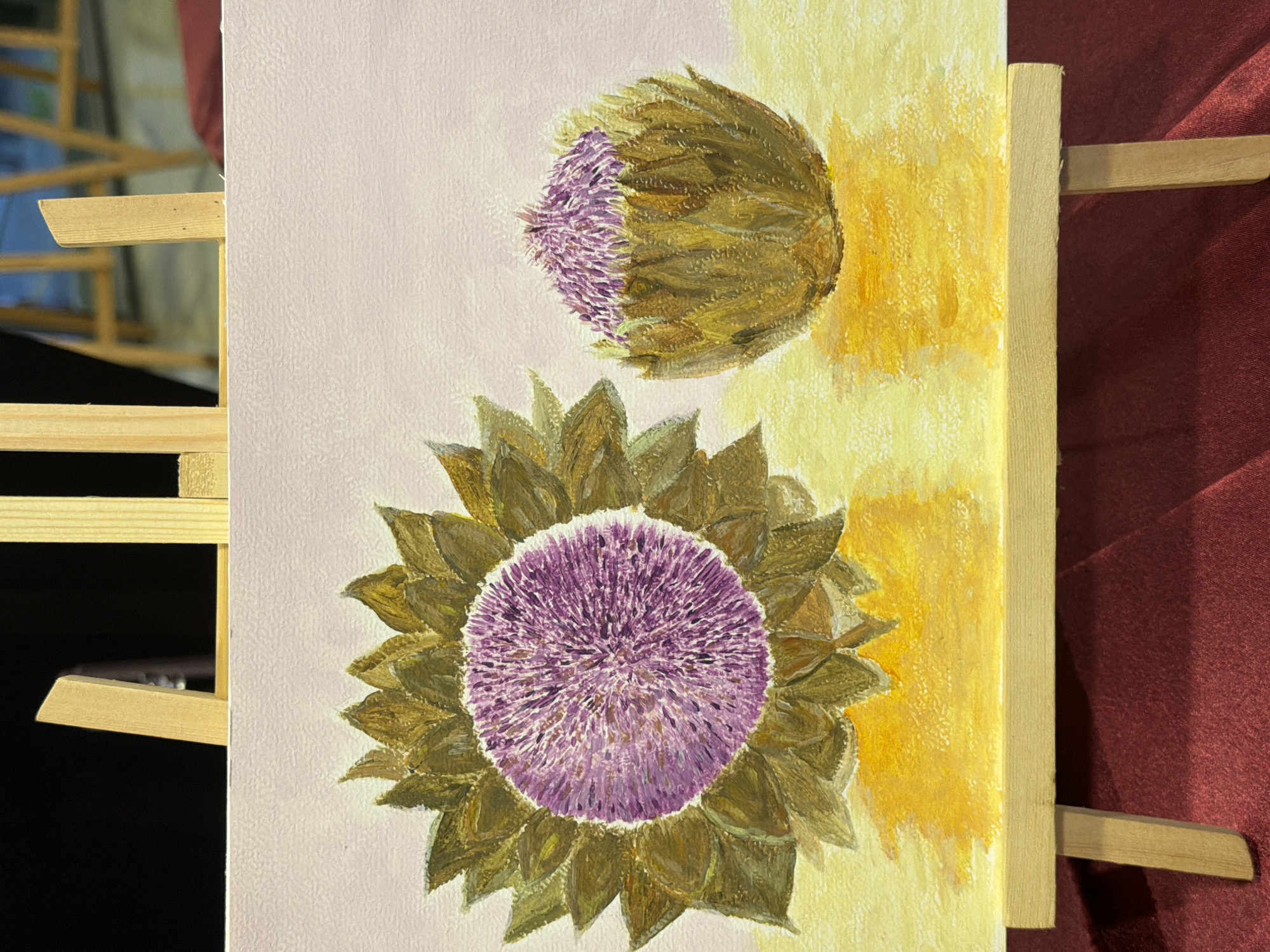 An oil painting on canvas depicts two artichokes on a yellow surface, against a pale pink background. The artichoke on the left is depicted as a full, blooming flower with a large purple center surrounded by spiky, green-brown petals. The artichoke on the right is smaller and appears to be in an earlier stage of development, with its purple florets peeking out from between protective green-brown leaves. Both artichokes are painted with visible brushstrokes, giving the texture of the canvas a prominent role in the artwork. The yellow surface beneath the artichokes is rendered with broad strokes of yellow and orange paint, suggesting light and shadow. The painting is displayed on a small wooden easel, which is positioned on a dark red textured surface, likely a tablecloth or fabric. The background behind the easel is dark, with some indistinct wooden structures visible. The painting is not located in Elche/Elx, Spain.