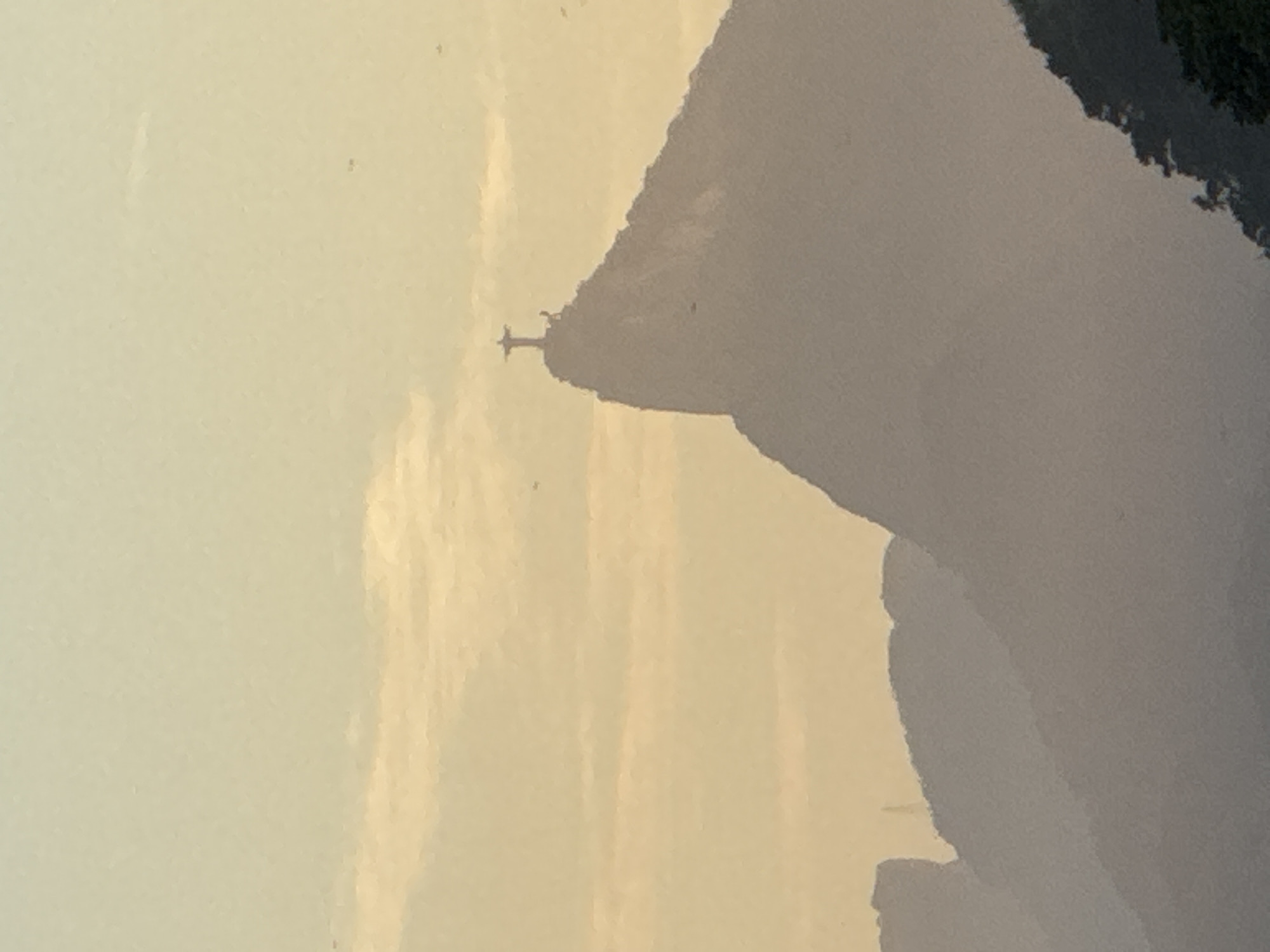 The image shows a silhouette of the Christ the Redeemer statue atop Corcovado Mountain in Rio de Janeiro, Brazil. The statue is visible against a soft, hazy sky with light clouds. The lower portion of the image displays a layered view of mountains fading into a misty atmosphere. The lighting suggests either sunrise or sunset, casting a warm, golden-yellow hue over the scene. No people, objects, or actions are discernable in detail due to the distance and atmospheric conditions.