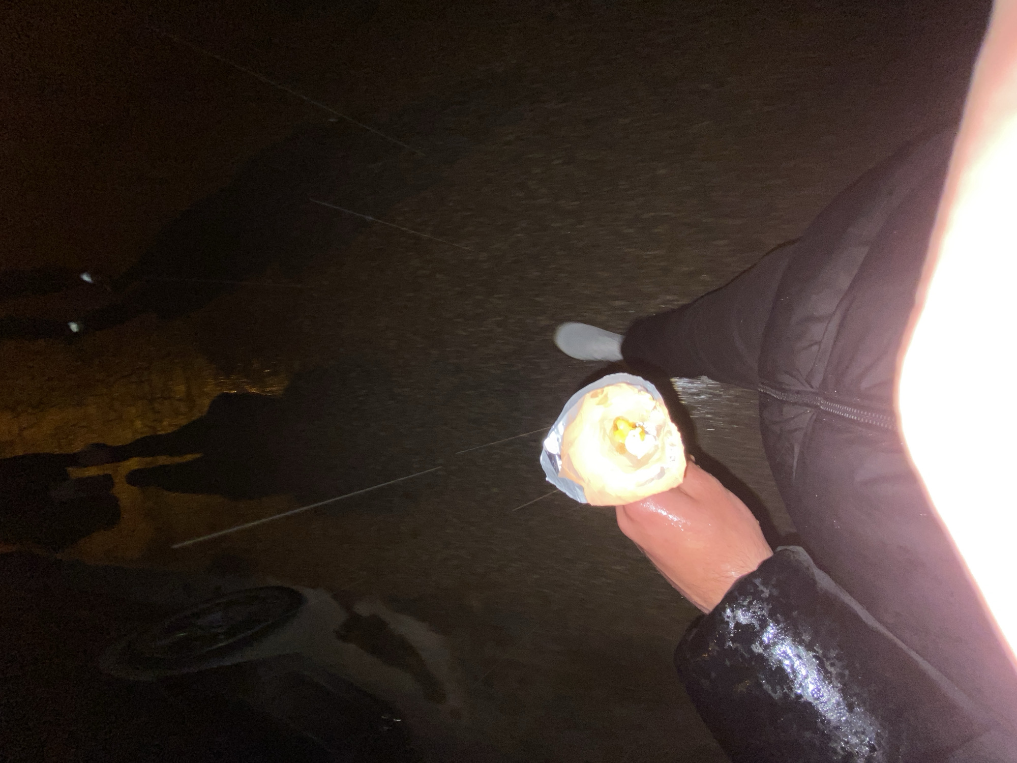 A person wearing black padded pants and a jacket holds a food item, likely a wrap or burrito, in aluminum foil. The ground appears to be wet and possibly asphalt, reflecting light. A white shoe is visible near the person. In the background, there are blurred reflections suggesting street lighting and possible parked vehicles. The overall lighting is poor, suggesting nighttime. Location information is unknown and cannot be verified from image content.