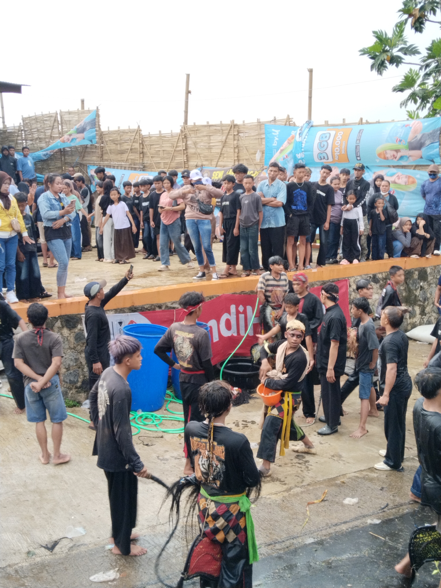 A medium, eye-level shot depicts a cultural ritual with a crowd observing, set in what appears to be Kuto, Indonesia. The foreground shows a concrete platform with numerous people, mostly young men, many wearing traditional attire. Some are in black outfits with red headbands, others in checkered patterns with green sashes. Some participants are barefoot, while others wear casual shoes. They are interacting, some possibly in a mock fighting style. A blue plastic barrel sits near the left side of the platform. A coiled green hose rests on the concrete.
Behind the platform, a crowd watches, standing on an elevated ledge. The crowd is diverse, with men, women, and children of varying ages. They are dressed in casual clothes, jeans, t-shirts, and some wearing head coverings. Some are holding phones, presumably taking photos or videos.
In the background, a bamboo-like structure serves as a makeshift fence, with advertisements attached to it. A tree is visible to the right. The sky is overcast, suggesting a cloudy day. The ground is wet, indicating recent rainfall or the ongoing nature of the event involves water.