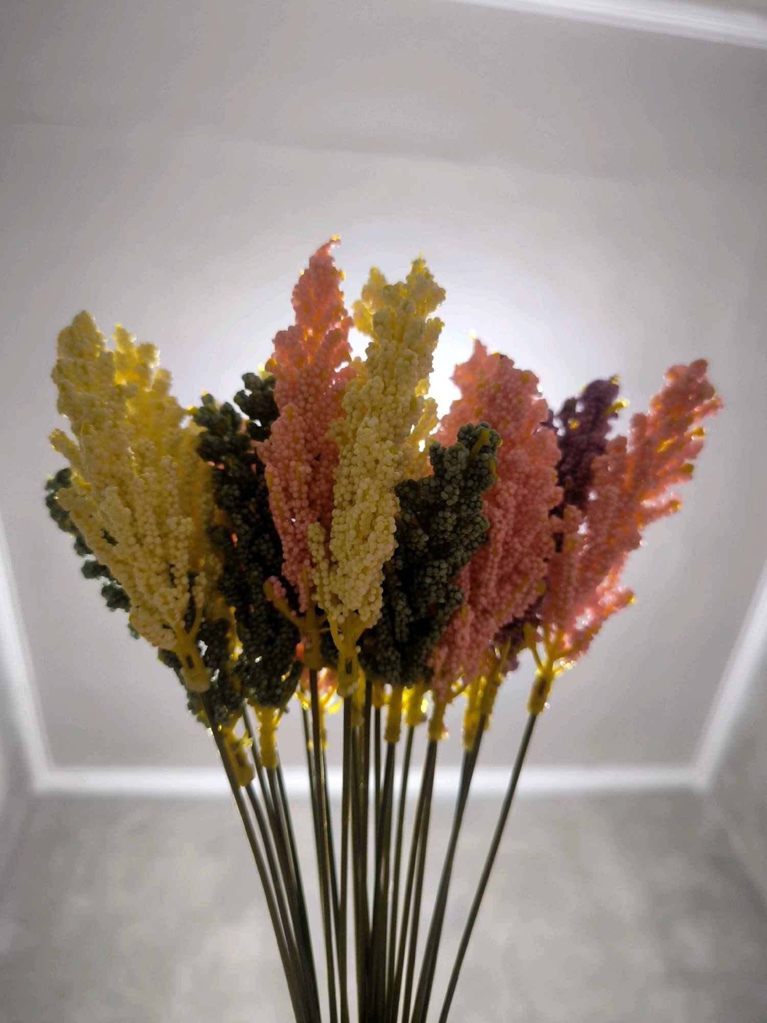 The image displays a close-up, low-angle shot of a bouquet of artificial flowers. The flowers are long and spiky, with a textured surface composed of small, bead-like elements. They are presented in a variety of colors, including yellow, pink, purple, and green. The bouquet is held together by thin, dark stems. The background is a bright, light-colored ceiling with subtle architectural details, possibly a recessed light fixture or coving. The lighting is soft and diffuse, suggesting an indoor setting. There are no people or discernible environmental elements that would indicate a specific location within Tabanan, Indonesia.