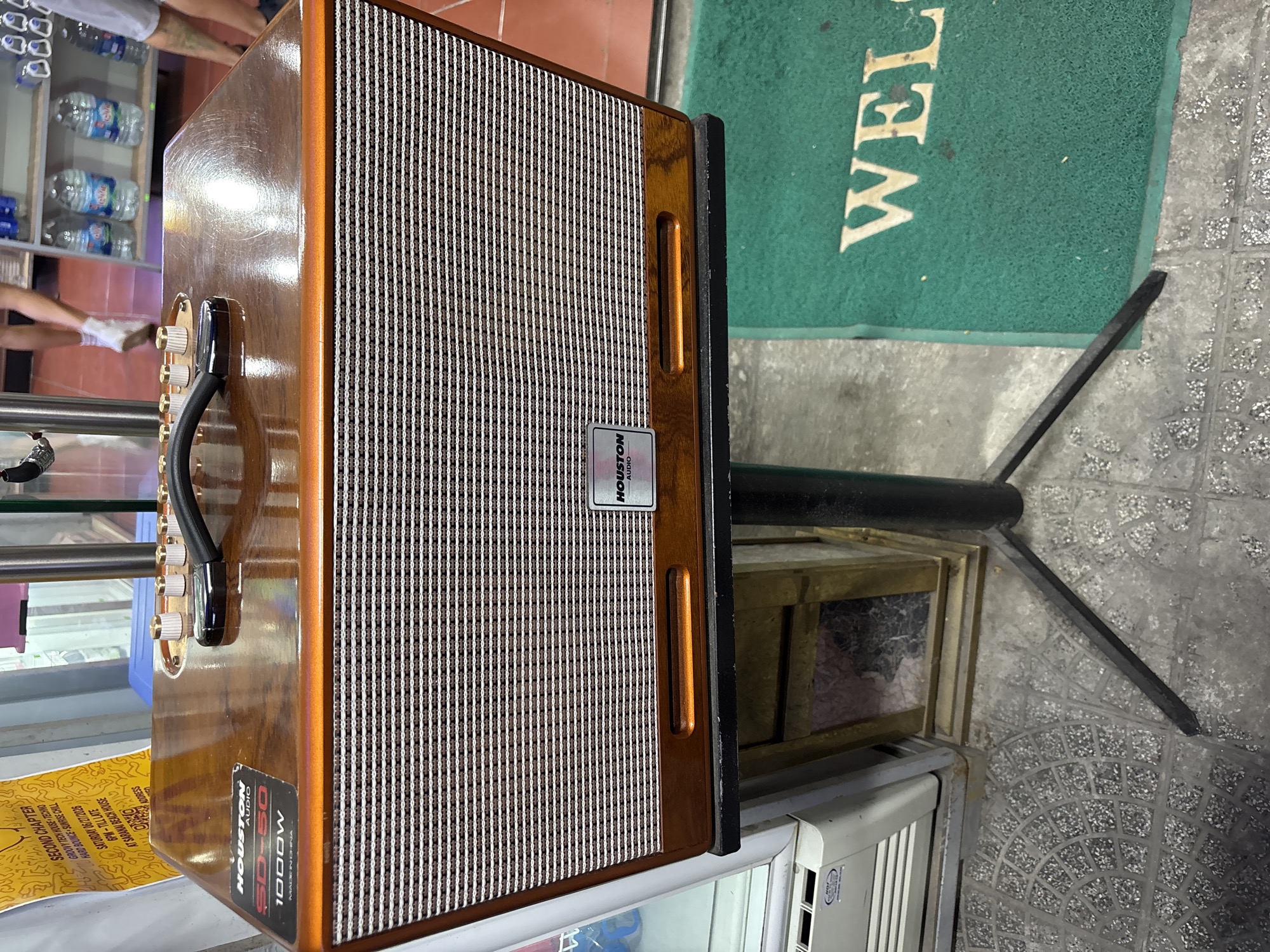 A glossy, wooden-finish speaker with a silver grille sits on a black tripod stand. The speaker has a chrome handle and three gold-colored knobs on its top surface. A sticker on the speaker's left side reads "HOUSTON AUDIO SD-50 1000W MADE IN CHINA". To the left of the speaker, a yellow poster is partially visible with Vietnamese text. In the background, shelves with plastic water bottles and an air conditioner unit are visible. A green welcome mat with the word "WELCOME" in white lettering is placed on the floor to the right of the tripod stand. The floor is tiled with a patterned design. Two people are partially visible in the background, their legs and lower bodies showing them walking. The scene appears to be an indoor shop or stall.