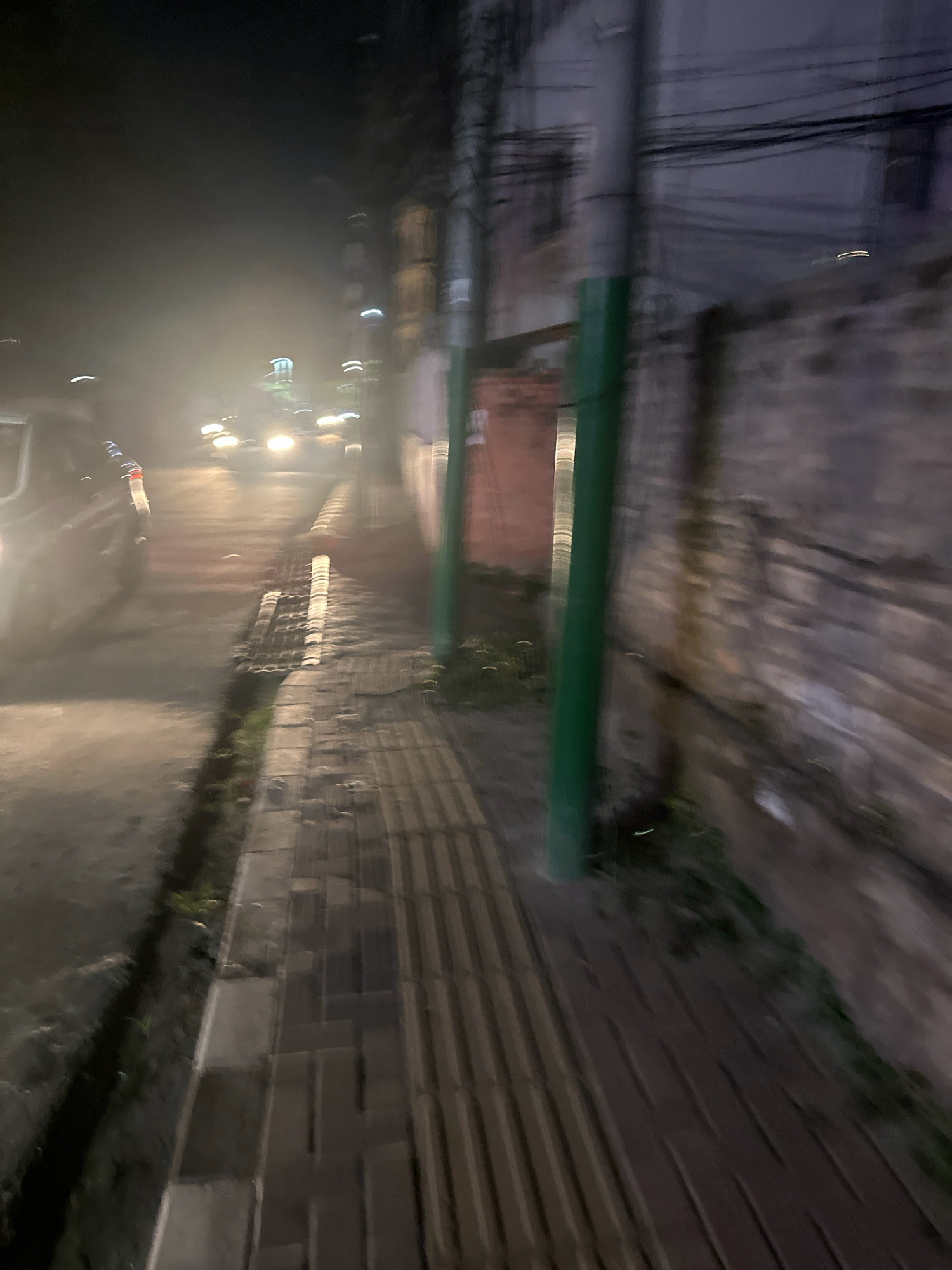 The image depicts a nighttime scene in Lalitpur, Nepal, characterized by motion blur. A vehicle with its headlights on is visible on a wet road, suggesting a recent rain or mist. The foreground features a brick-paved sidewalk with raised tactile paving for visually impaired pedestrians. A green pole stands vertically on the sidewalk, and behind it, a brick wall with some moss or vegetation growth is present. Further back, buildings and overhead electrical wires are discernible. The overall impression is of a street scene in a city at night, with limited visibility due to the blur and possibly atmospheric conditions.