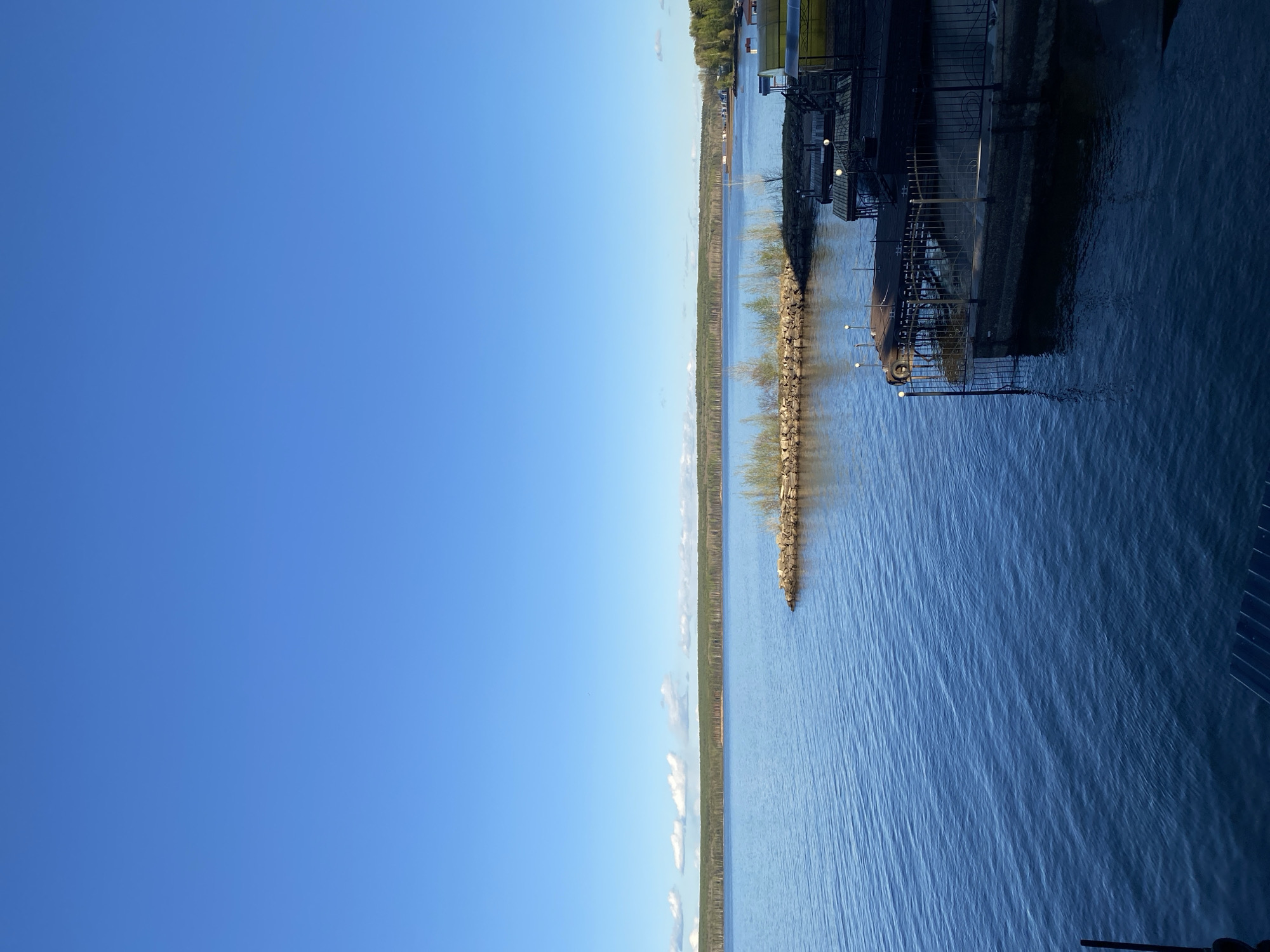 The image depicts a waterfront scene in Chaikovsky, Russia, featuring a view of the Kama River. A rock jetty extends into the river, positioned between a tree-lined shore and a dark structure on the right side of the frame, potentially a restaurant or dock. The sky is clear and blue. The water is visible and appears calm, reflecting the sunlight. The photograph's angle is tilted.