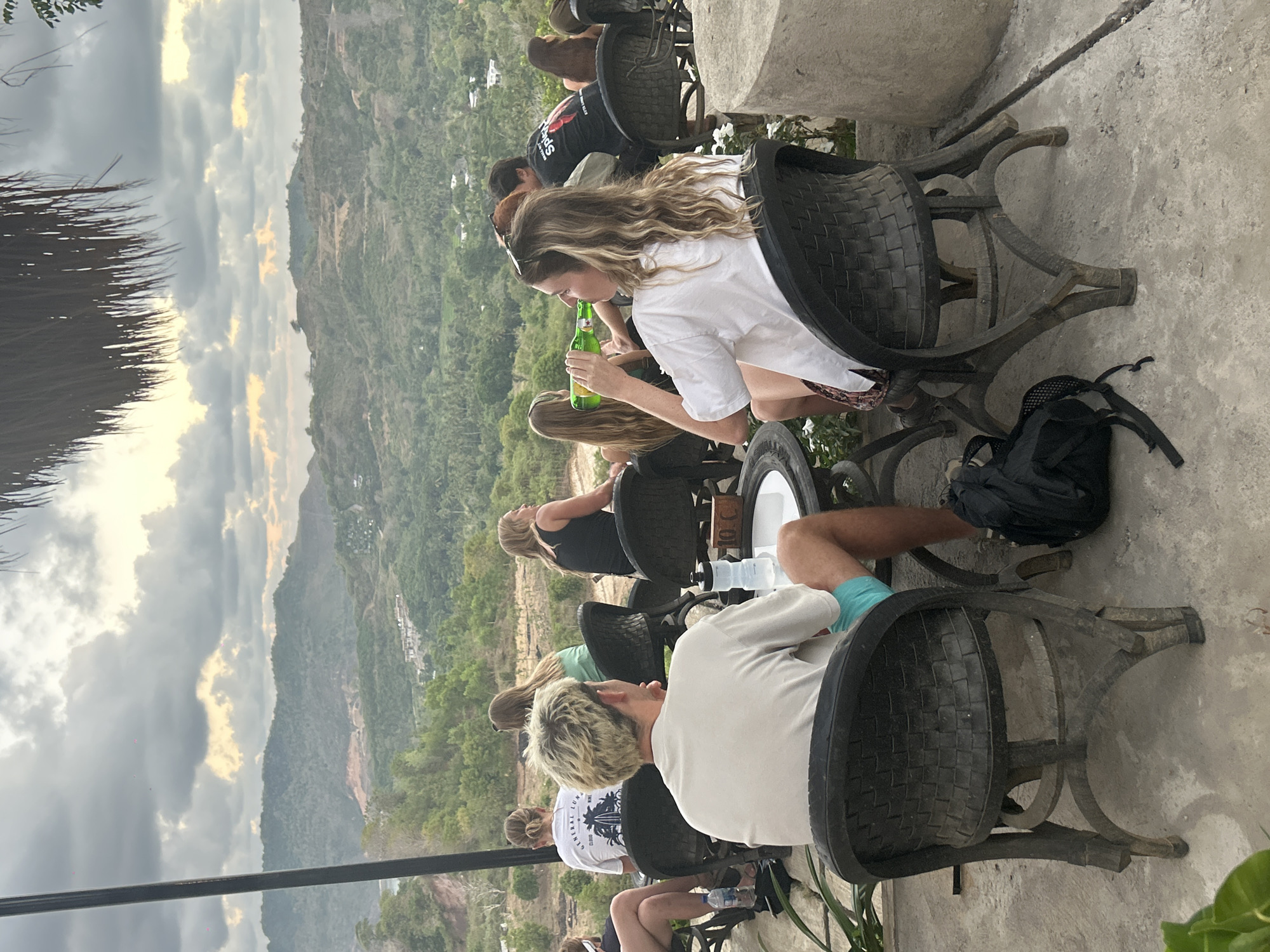 The image captures an outdoor seating area at a venue overlooking a lush, hilly landscape under a cloudy sky. In the foreground, several people are seated at tables, engaged in conversation and enjoying drinks. A woman in the right foreground is drinking from a green bottle. A man in the center foreground is facing away from the camera, wearing a light-colored t-shirt and turquoise shorts. The chairs appear to be made of woven black material with unique curved legs. In the middle ground, other patrons are visible, some with their backs to the camera. The background reveals a vista of rolling hills covered in dense greenery, with scattered buildings and evidence of construction or terraced farming. The overall atmosphere suggests a relaxed setting, possibly a cafe or restaurant, in a tropical or subtropical location like Kuta, Indonesia, known for its scenic beauty and outdoor dining options.