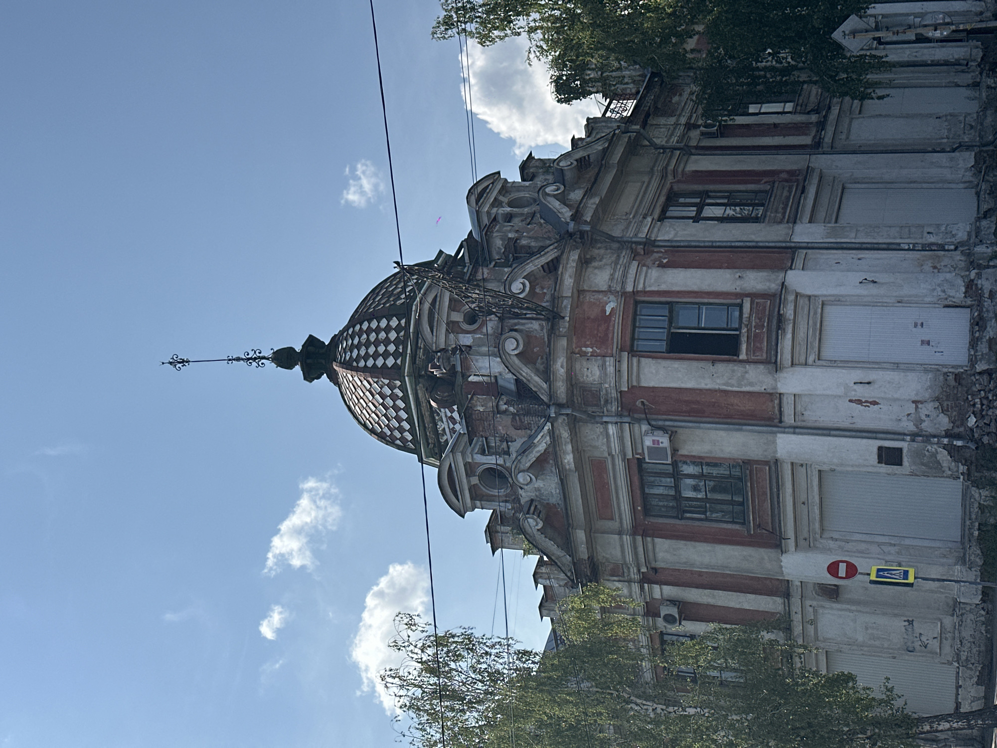 The image shows a weathered, ornate building with a domed roof topped by a spire. The building's facade is painted in alternating vertical stripes of white and faded red, with areas of peeling paint and visible brickwork indicating disrepair. The windows have dark frames, and some appear to be boarded up or covered. There are air conditioning units mounted on the facade.
In front of the building, there are two traffic signs: one circular red sign with a white horizontal bar, and below it, a diamond-shaped yellow sign with a pedestrian symbol and an arrow. Several trees with green foliage are visible on either side of the building, partially obscuring the view. Overhead, power lines stretch across the frame against a blue sky with scattered white clouds. The building is situated in the city of Biysk, Russia, as indicated by the presence of the pedestrian traffic sign, which is common in Russia. The overall scene depicts a historic, somewhat neglected urban building.