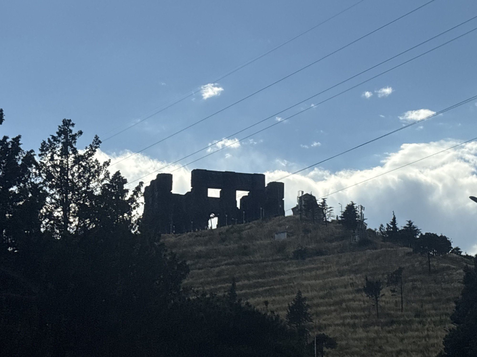 The image shows a silhouette of a ruined stone structure on a grassy hillside under a partly cloudy blue sky. The structure appears to be a large, rectangular building with multiple openings, possibly windows or doorways. It is silhouetted against the sky, making its details difficult to discern. The hillside is covered in dry grass and scattered trees, with terraced lines suggesting agricultural use or erosion control. Several utility poles with wires are visible stretching across the sky, and there are some smaller structures and trees on the hillside near the main ruin. The overall impression is one of historical ruins set against a natural landscape. The lighting suggests it is daytime, possibly late afternoon due to the angle of the sun and the shadows. No people are visible.