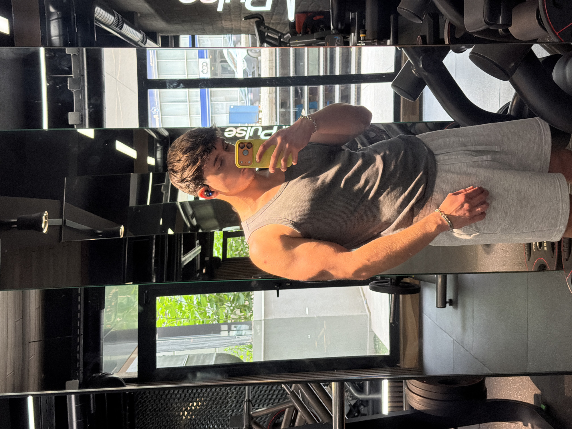 The image shows a man in a gym, taking a selfie in a mirror. The reflection in the mirror shows buildings outside, indicating an urban setting. "PULSE" is visible in the reflection, suggesting the gym name. The gym appears to be located in Sofia, Bulgaria, as evidenced by the presence of a "Volkswagen" dealership and buildings typical of the city's architecture in the background. The man is wearing a tank top and shorts, and has earbuds in his ear. He is holding a yellow phone. Gym equipment is visible in the foreground and background.