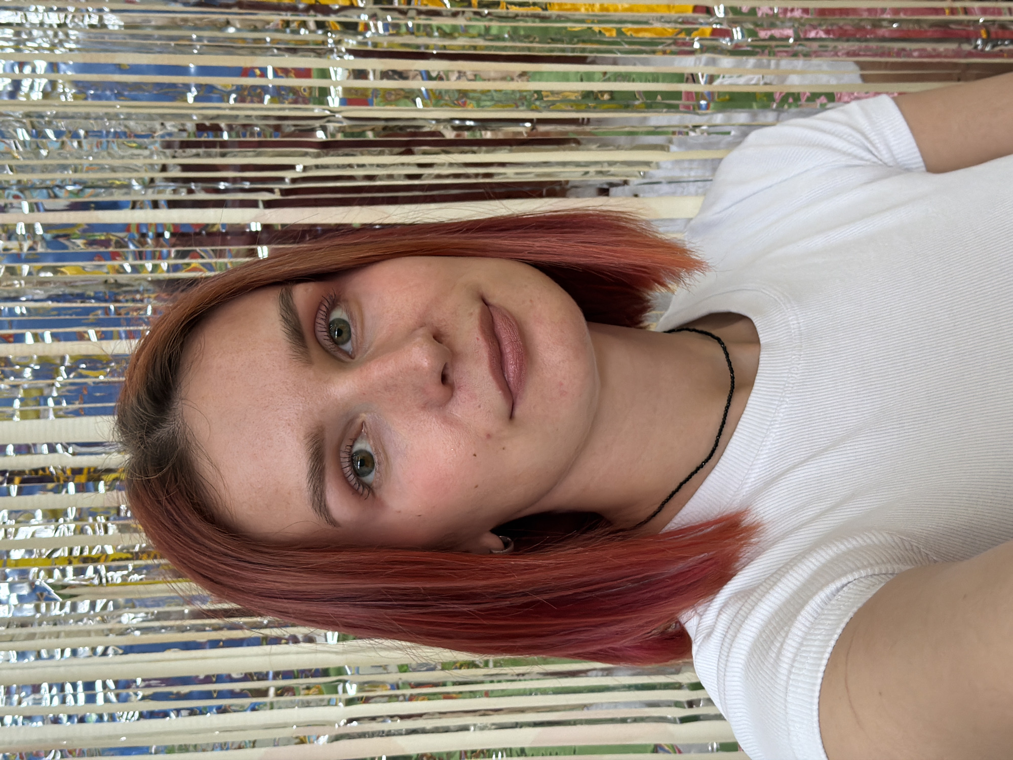 A close-up, eye-level portrait depicts a young woman with short, reddish-pink hair, fair skin, and light green eyes. She is wearing a white t-shirt and a black beaded necklace. The background consists of a shiny, metallic, horizontally-striped surface, likely a party decoration. There's no information available to suggest the location in the city of Chaikovsky, Russia. The subject is looking directly at the camera with a subtle smile.