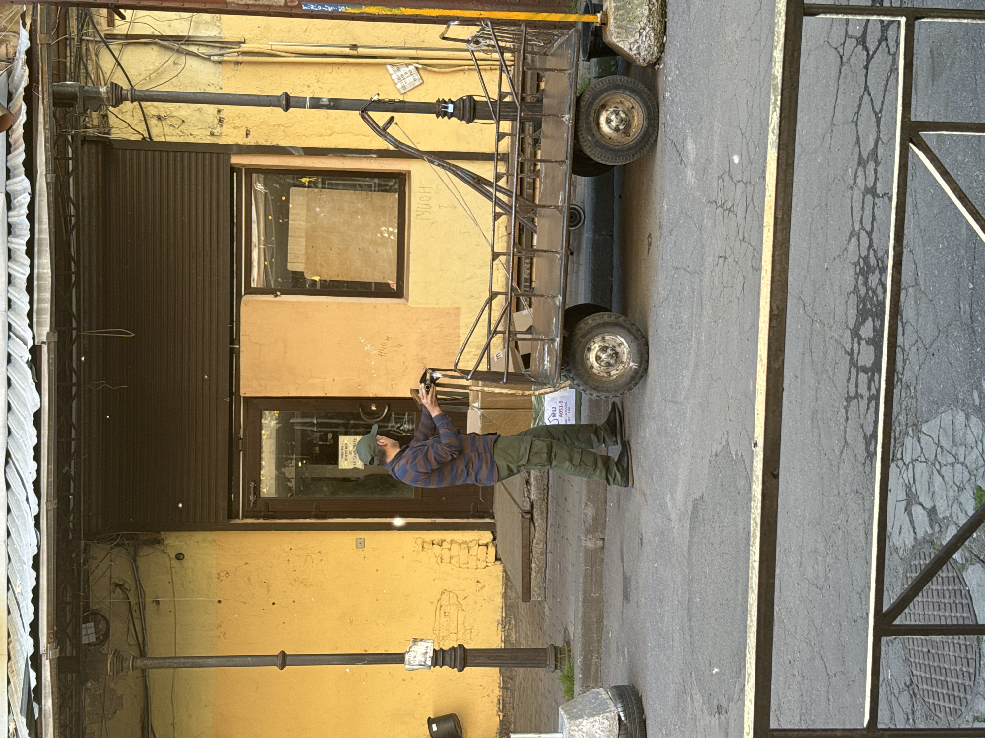 A man in a green baseball cap, a blue and brown striped long-sleeved shirt, and green cargo pants is pushing a large, rusty, four-wheeled metal cart on a cracked asphalt sidewalk. The cart has a raised platform with a metal frame and appears to be empty, except for a small piece of metal structure attached to its center. The man is positioned to the left of the cart, with his hands on the handles, and he is facing away from the camera, towards the right.
Behind the man and the cart, there is a building with a bright yellow facade. The building has a dark brown metal roll-up door on the right side, partially covering an entrance. To the left of the roll-up door is a dark brown door frame with glass, through which a person can be dimly seen. A window with a dark frame is situated above and to the left of the entrance. The building's facade is weathered and shows signs of wear and tear, with some brickwork visible in places.
The scene is lit by natural daylight, casting shadows on the ground. There are several elements in the environment: a black metal lamp post with a sign attached to it is to the left of the man. A concrete block with a tire inside is located on the sidewalk to the far left. Another lamp post, also black, is to the right of the cart. A yellow metal pole is visible on the far right of the image. A metal fence runs across the foreground of the image, obscuring the lower portion of the scene.
The location is identified as being in Saint Petersburg, Russia, based on the provided context.