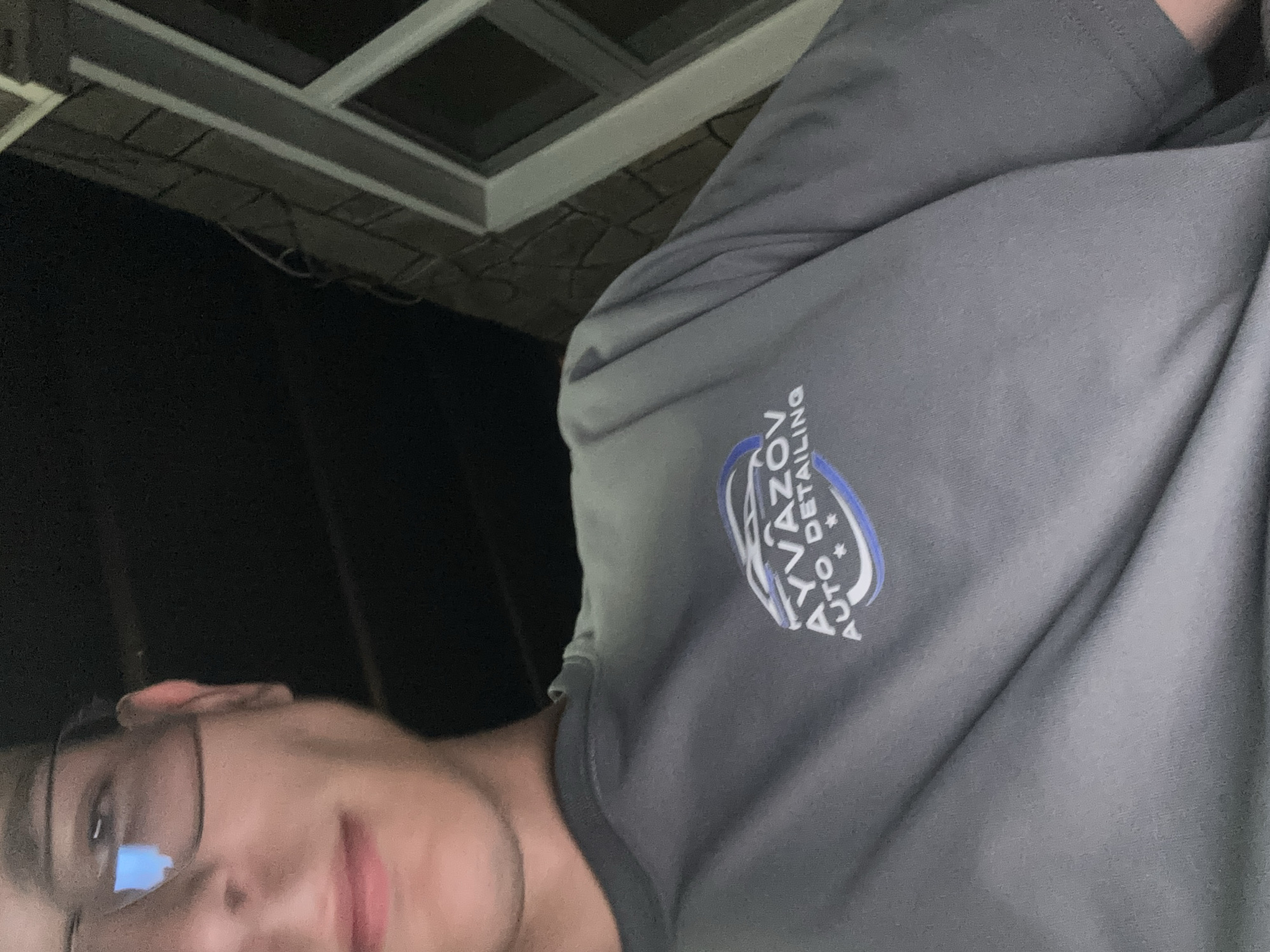 A close-up, low-angle, forward-facing shot shows the upper body and head of a young person wearing glasses and a grey t-shirt with a blue and white logo that reads "AYVAZOV AUTO DETAILING." The person's face is partially visible, with their mouth open as if speaking or smiling. The background is dark, with a hint of a building with brick siding and a window on the right side. The image appears to have been taken at night. The city of Nedelino, Bulgaria, is not depicted in this image.