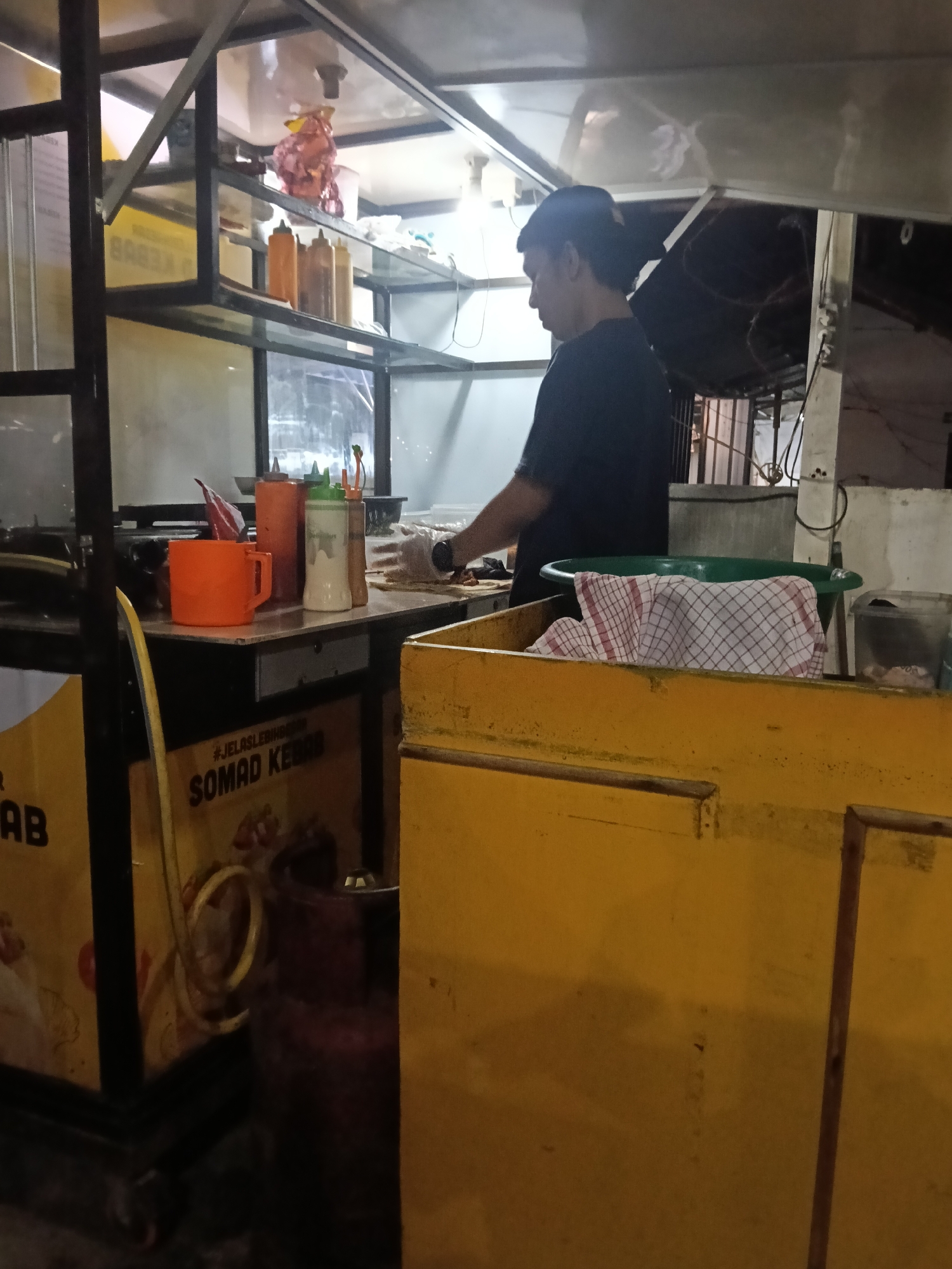 The image depicts a food stall in Lambheu, Indonesia. A man, presumably the vendor, is seen working behind the counter. The stall appears to be preparing or serving kebabs, as indicated by the text "#JELASLEBIHBERSIH SOMAD KEBAB" visible on the side of the stall. Various containers, including sauce bottles and a red cup, are arranged on the counter. A gas cylinder is positioned near the stall. The lighting suggests it is nighttime or dusk.