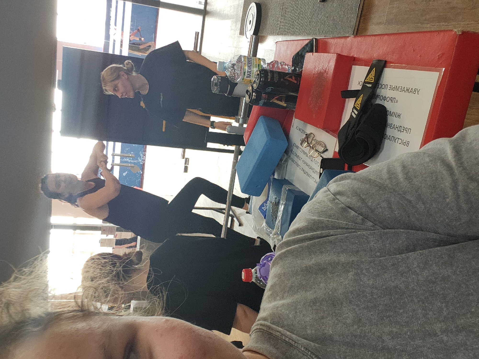 The image shows an indoor space, possibly a gym, with several people present. A woman in black workout attire is stretching, and another woman in black stands nearby, seemingly observing. In the foreground, a person in a gray garment is positioned close to the camera. Gym equipment, water bottles, and signage with Cyrillic text are scattered around. The location is unidentified.